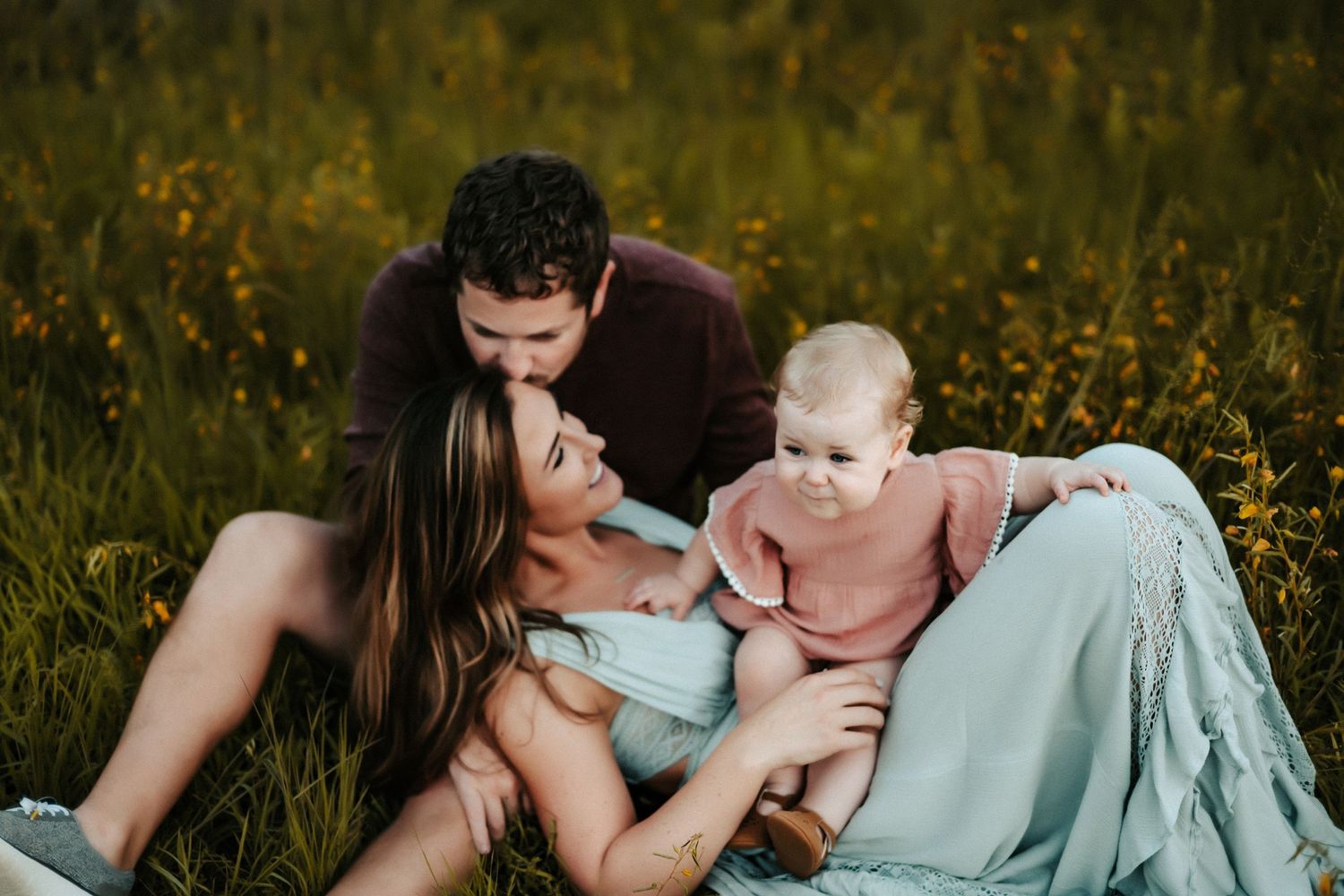 Family of three sitting together in a wildflower meadow during golden hour wearing casual clothing.