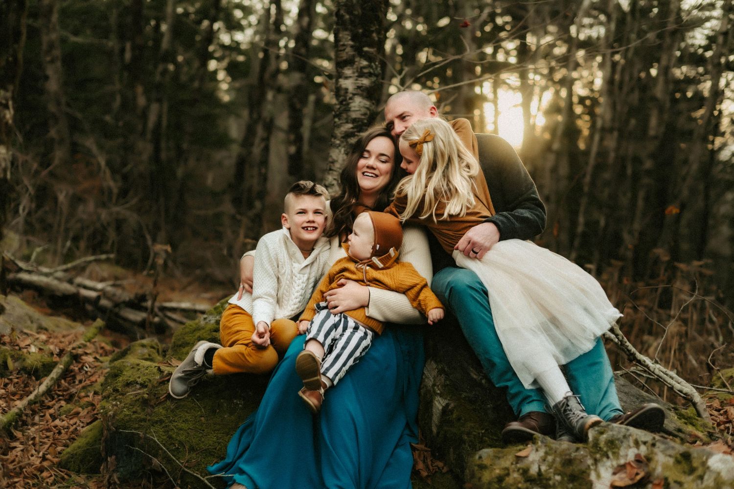 A loving family sits together on fallen logs in an autumn forest at sunset with golden sunlight filtering through trees.