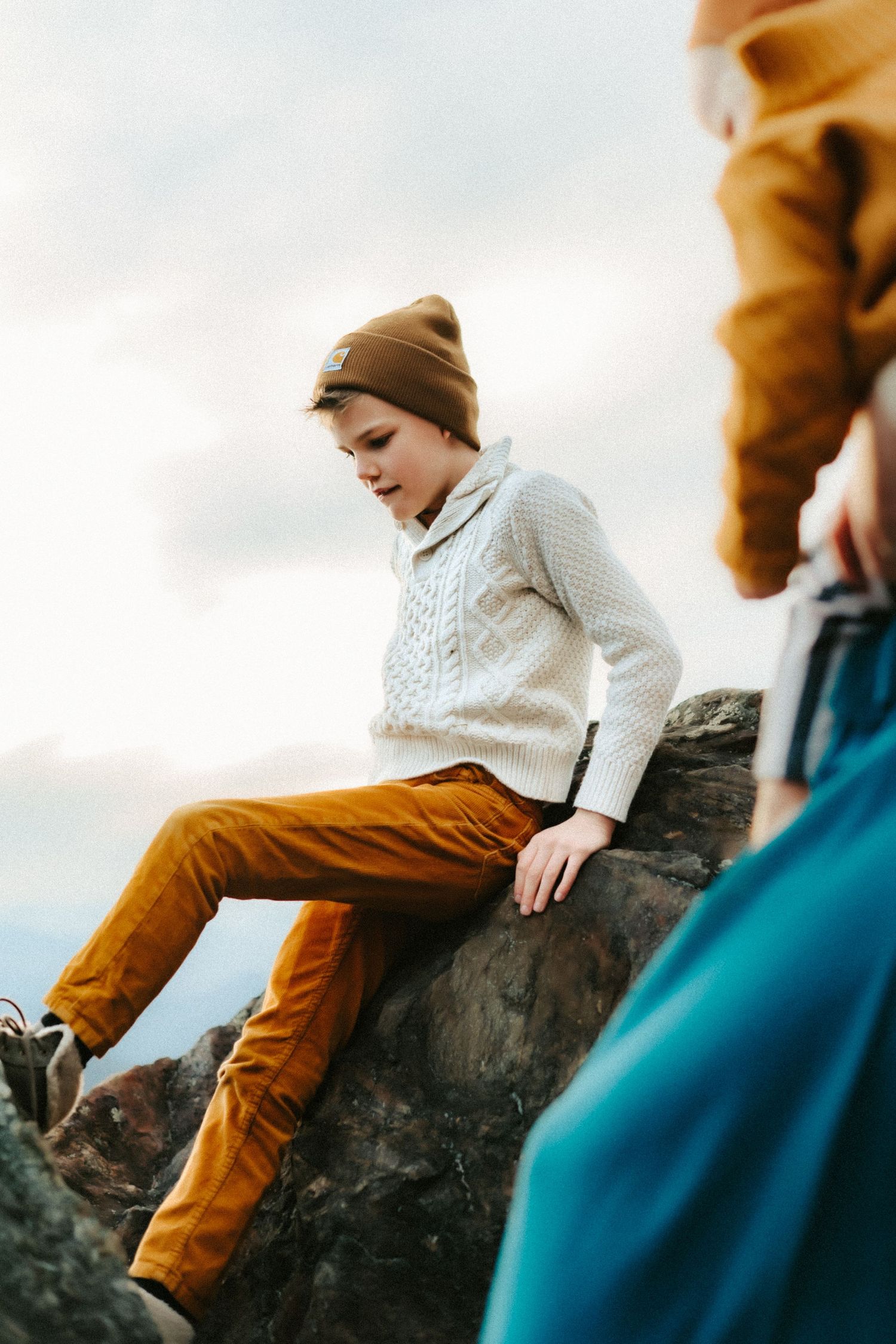 Person in beige beanie and cream sweater sitting thoughtfully on rock formation against cloudy sky.