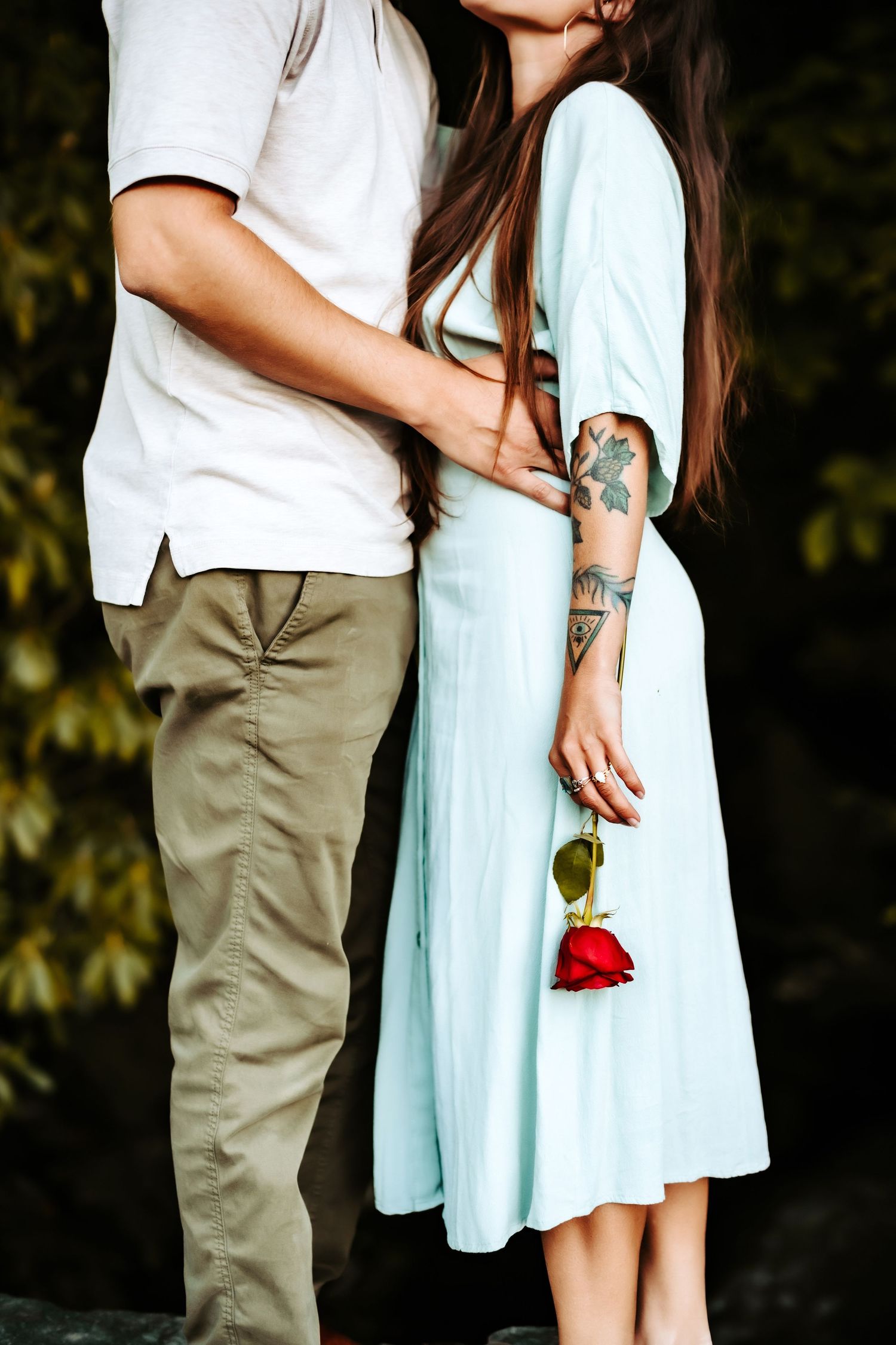 A romantic couple embracing in mint and white outfits while holding a red rose against a leafy background.