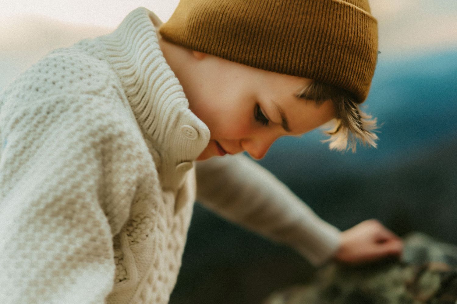 Person in a brown beanie and cozy white sweater bends down to examine something outdoors on a cloudy day.