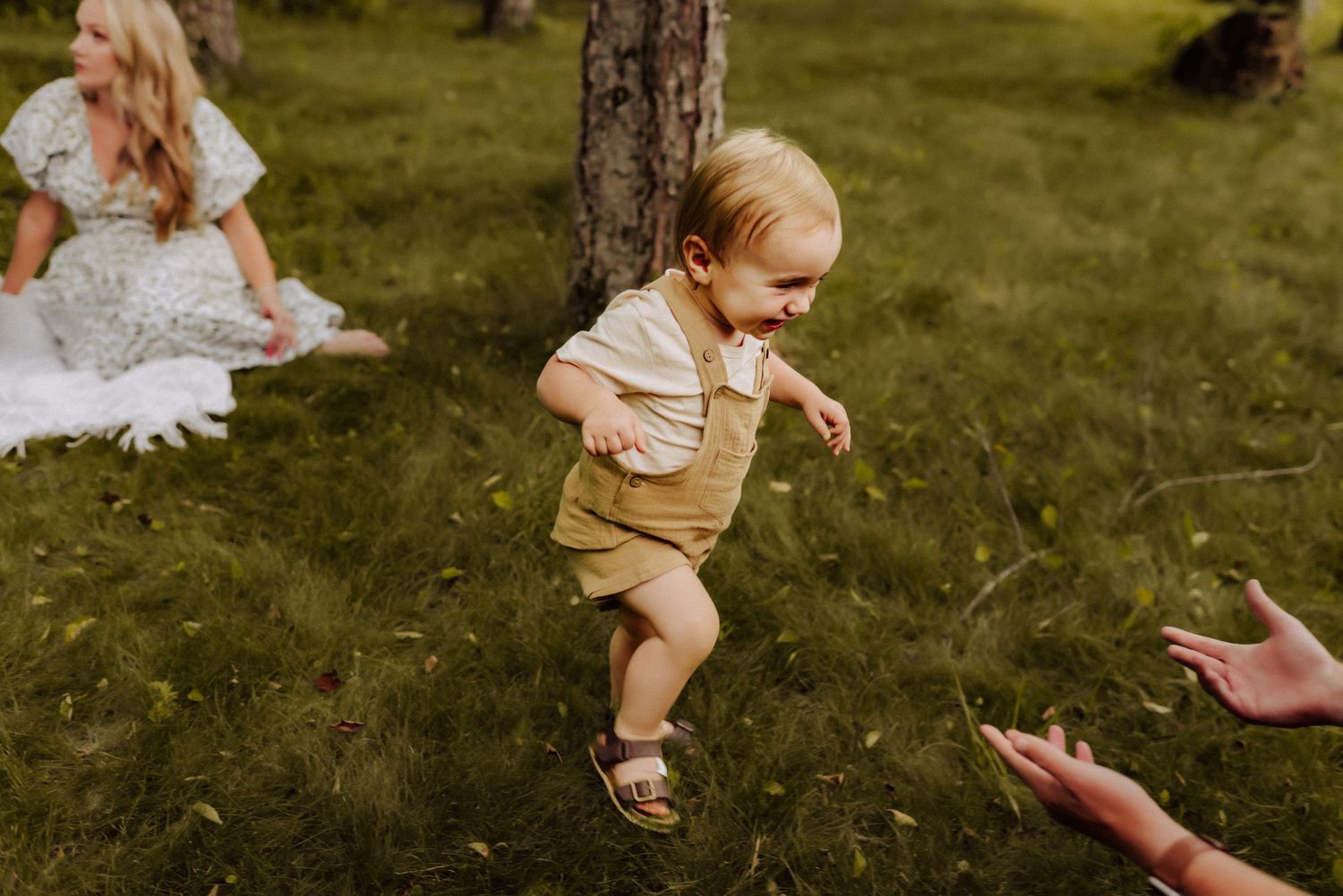 Child in beige outfit running across grass during outdoor picnic as hands reach out in greeting.