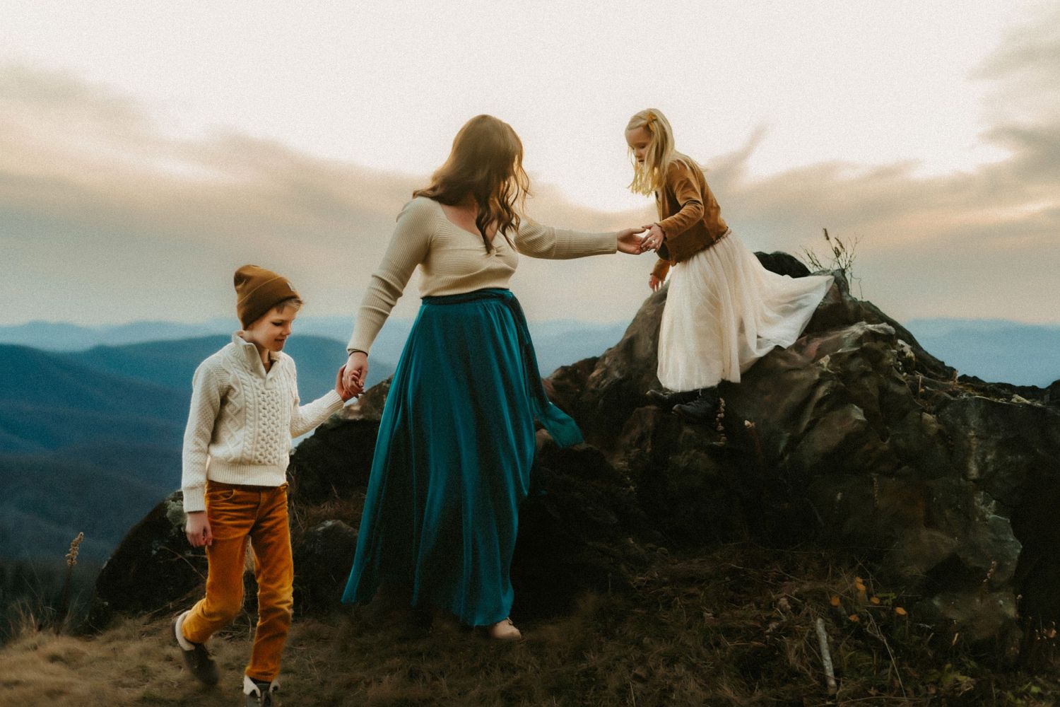 People in casual clothing stand together on a mountain summit at sunset overlooking a vast mountain landscape.