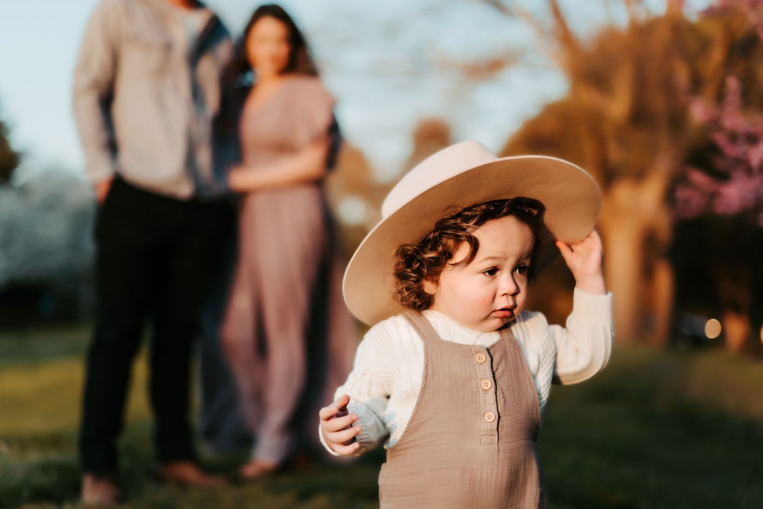 A toddler in a tan outfit and wide-brim hat explores outdoors during golden hour with blurred people in the background.