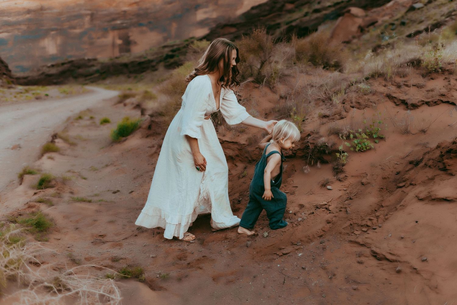 A woman in a white dress guides a small child along a rocky desert trail surrounded by red sandstone formations.