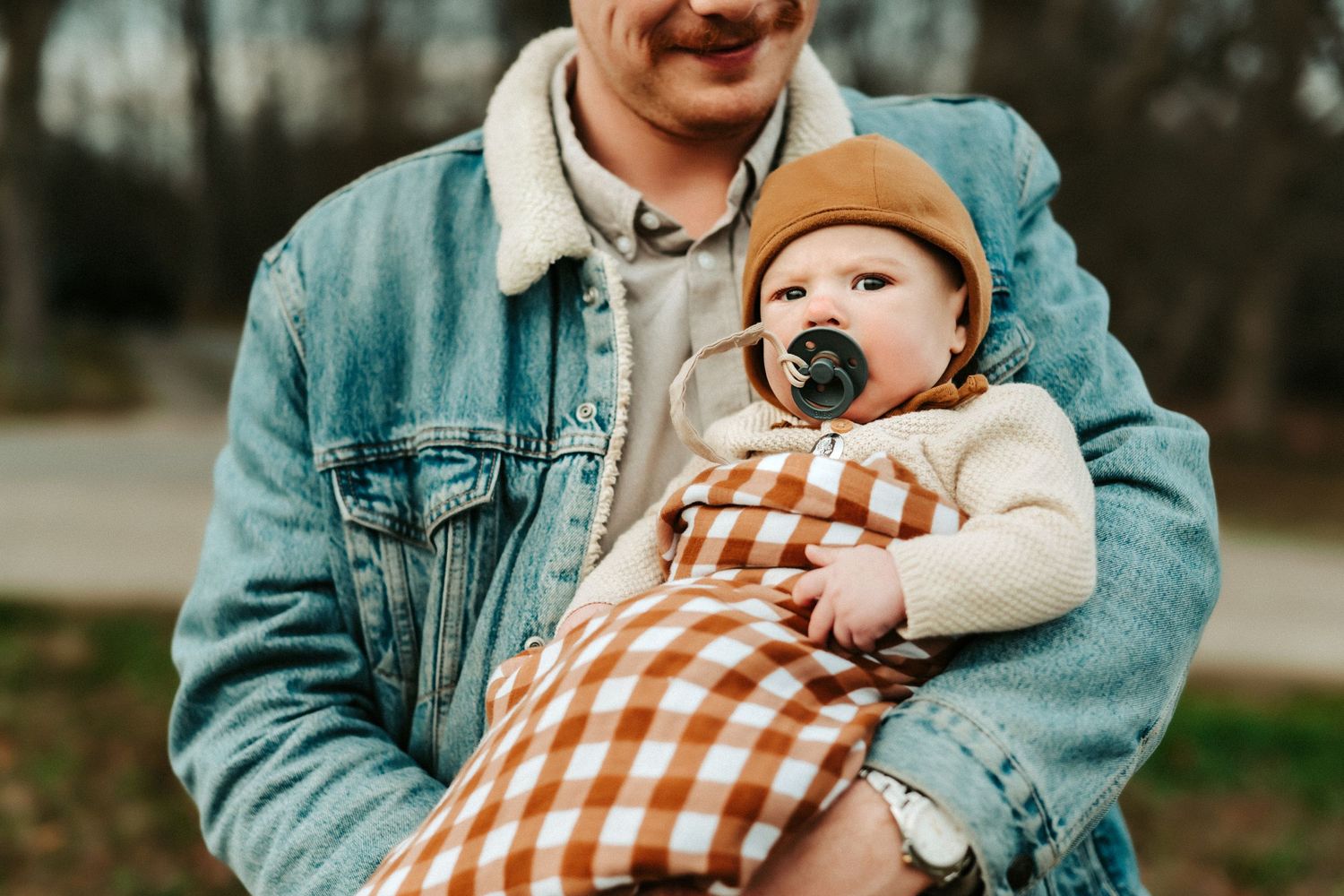 Baby wearing an orange beanie and gingham blanket is held in someone's arms wearing a denim jacket.