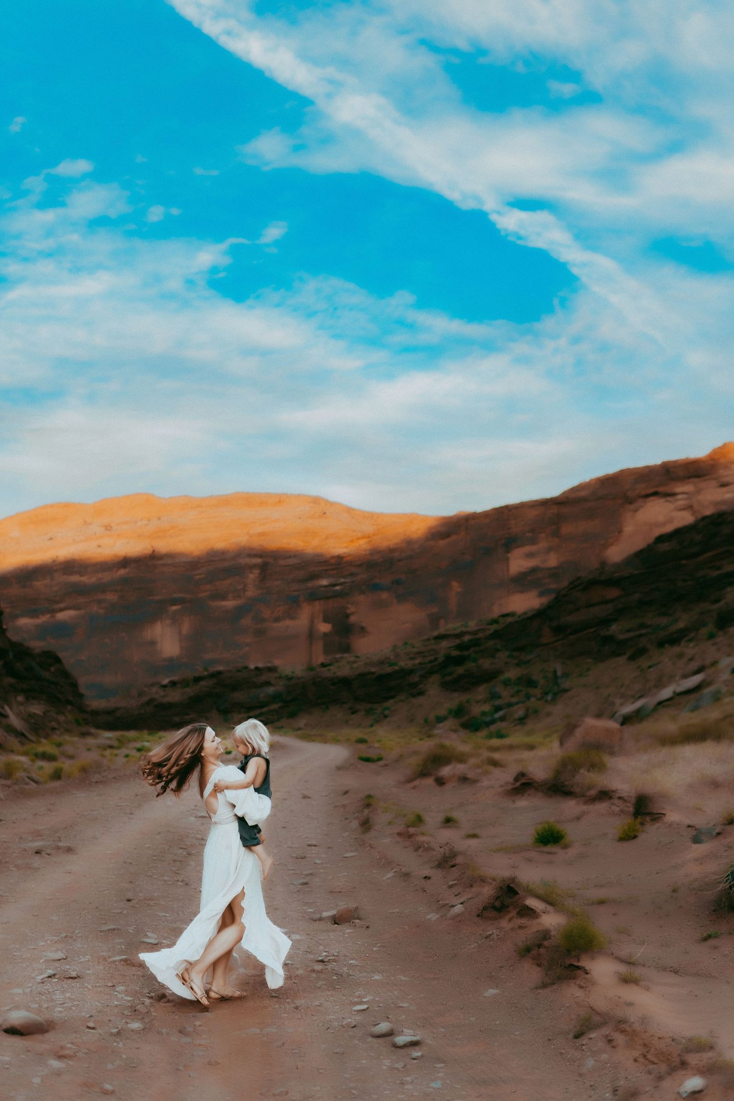 A couple shares a romantic moment in flowing white attire against a dramatic desert landscape under bright blue skies.