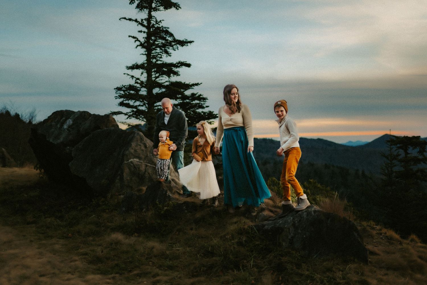 Group stands on mountain peak at sunset with evergreen tree silhouetted against dramatic sky in background.