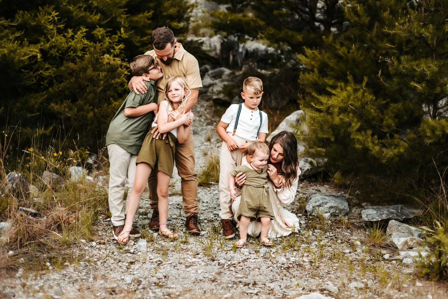 A group in coordinated neutral outfits shares joyful laughter together on a rocky outdoor terrain surrounded by evergreens.