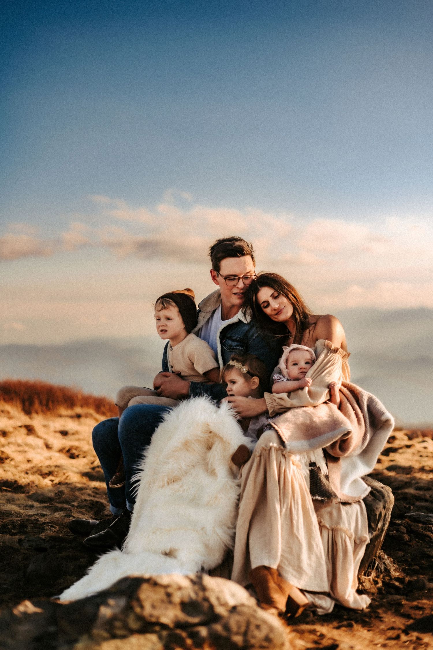 A family sits together on a rocky hilltop at sunset with their fluffy white dog, creating a warm and intimate moment.
