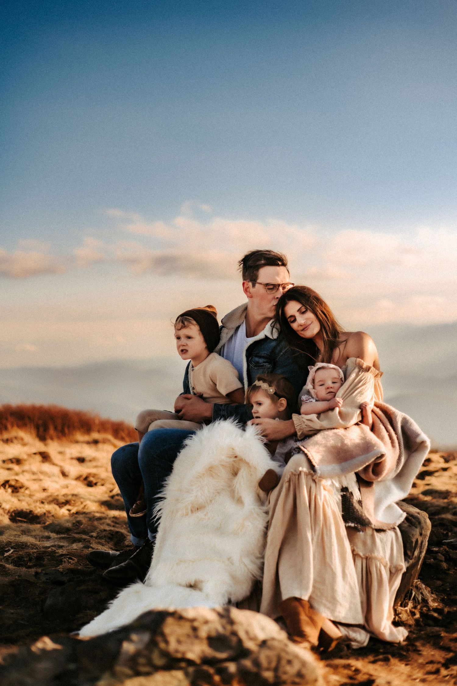 Group sits together with fluffy white dog on scenic hilltop during golden hour sunset.