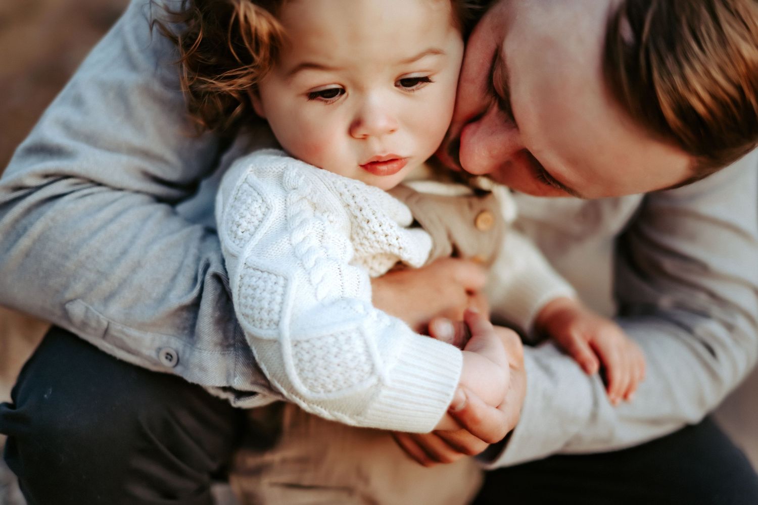 A small child in a white sweater snuggles close to a parent in a tender moment on gray bedding.