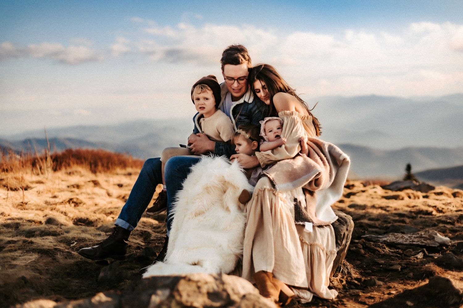 A group sits together with their white dog on a mountaintop during golden hour with misty peaks in the background.