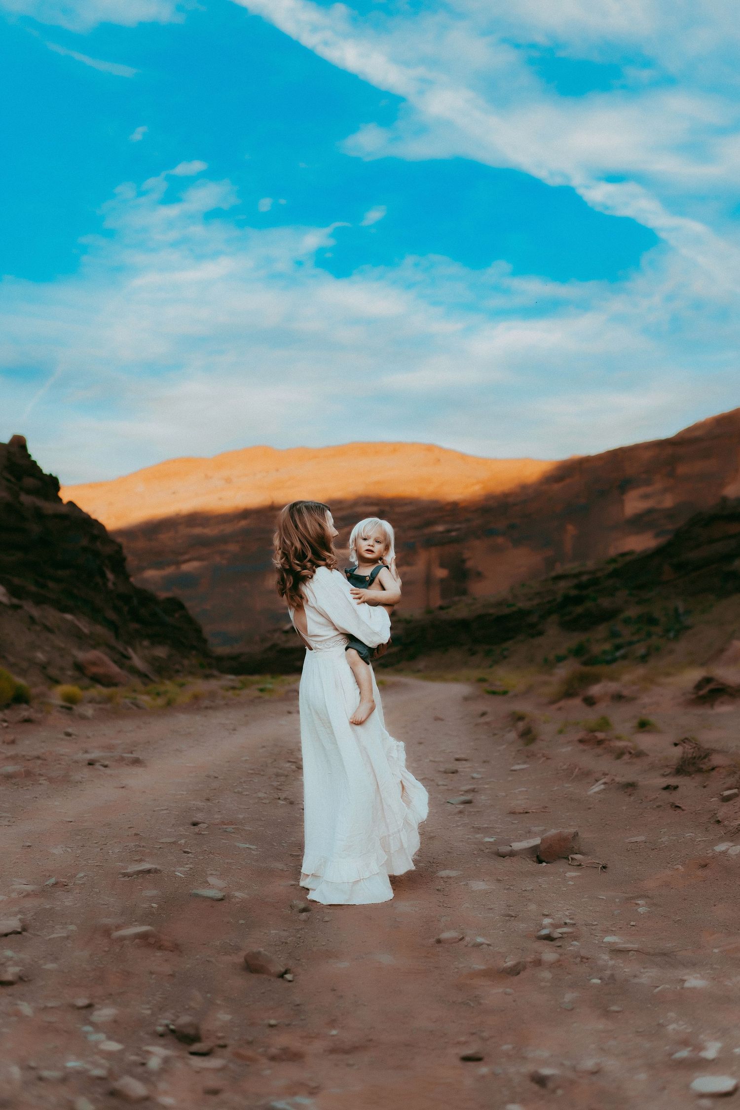 Figure in a flowing white dress stands on a desert path with golden hills and blue skies in the background.