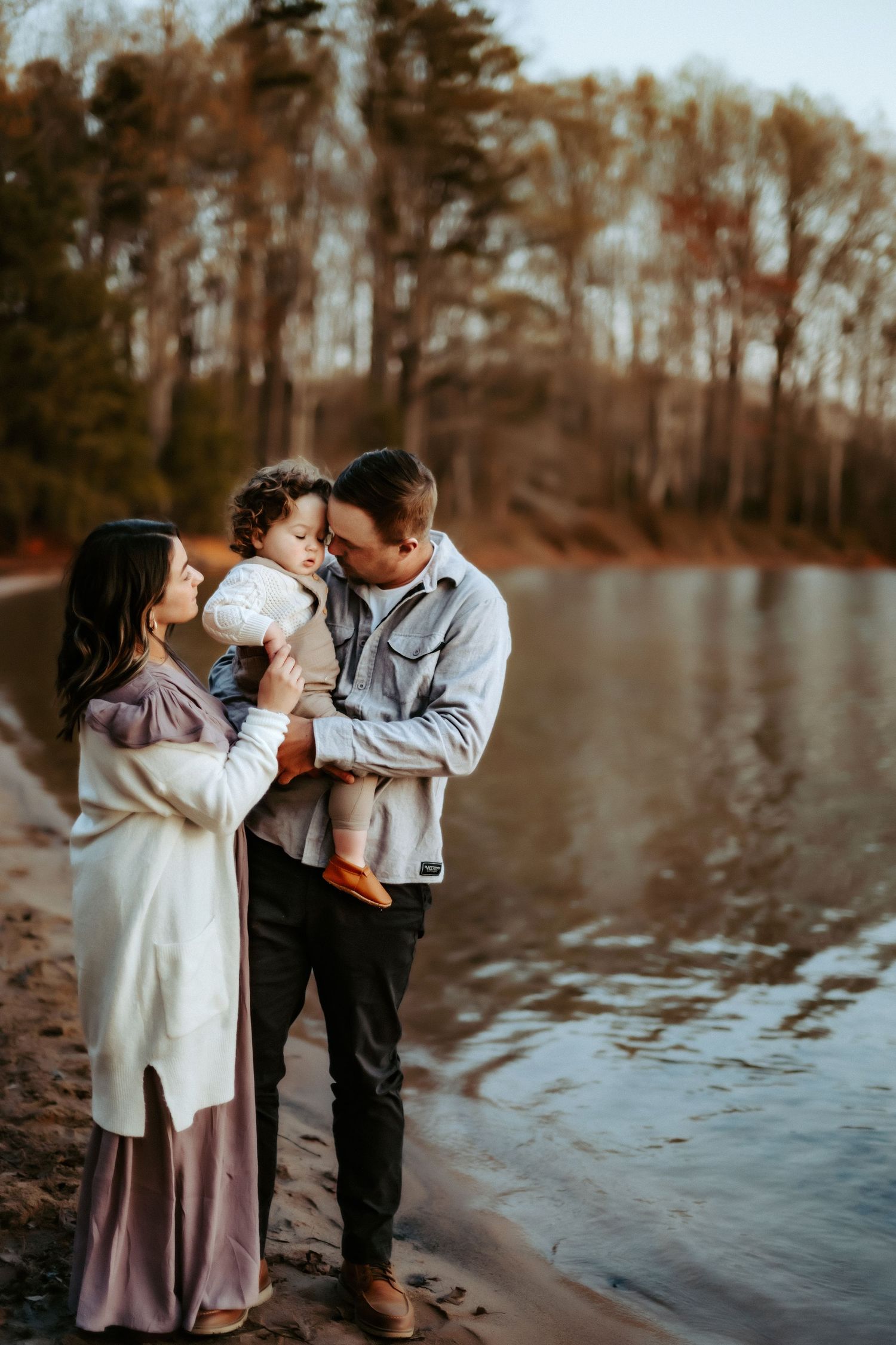Family shares tender moment together at sunset by a serene lake with autumn trees in background.