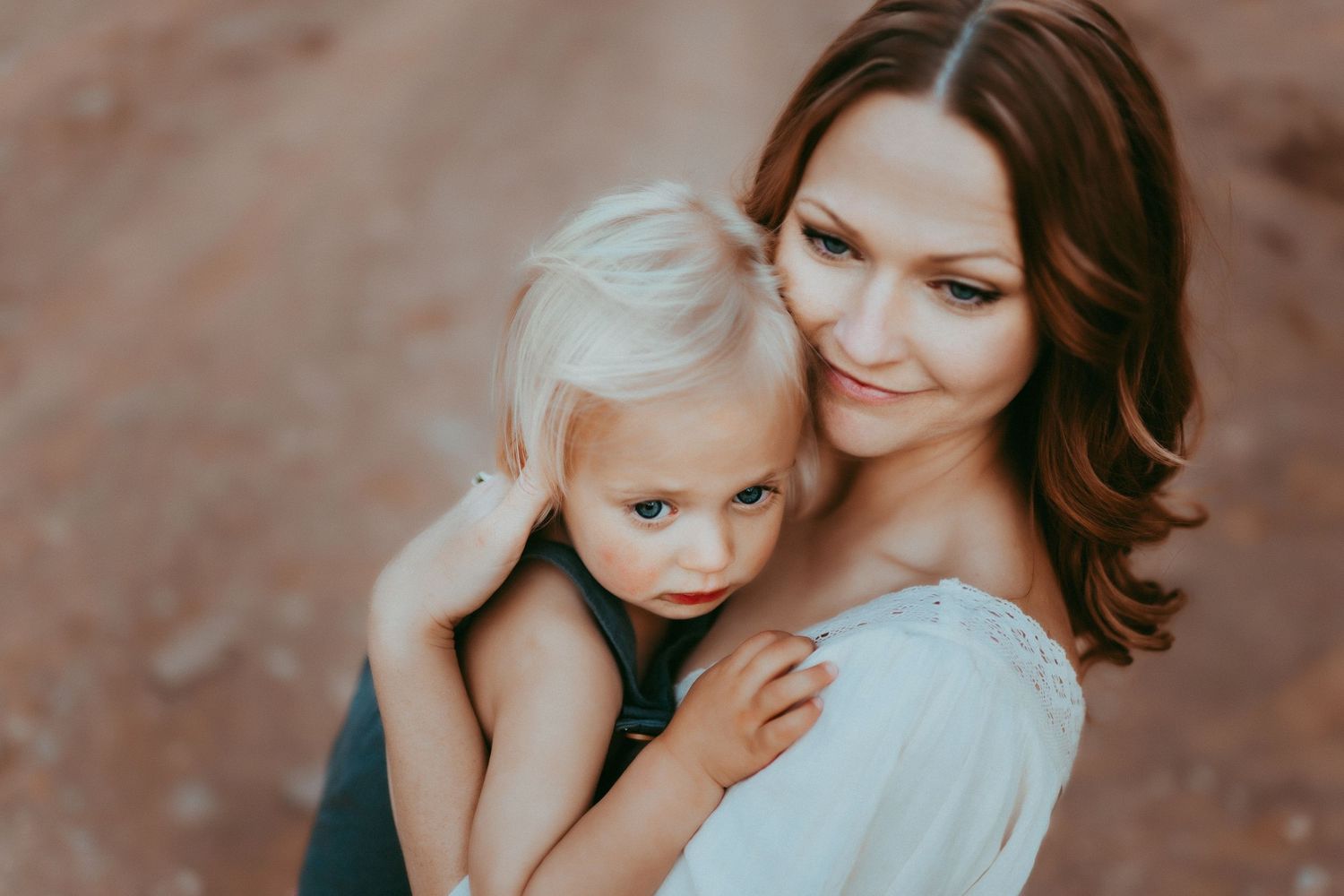A loving parent in a white top embraces their blonde-haired child outdoors against an earthy background.