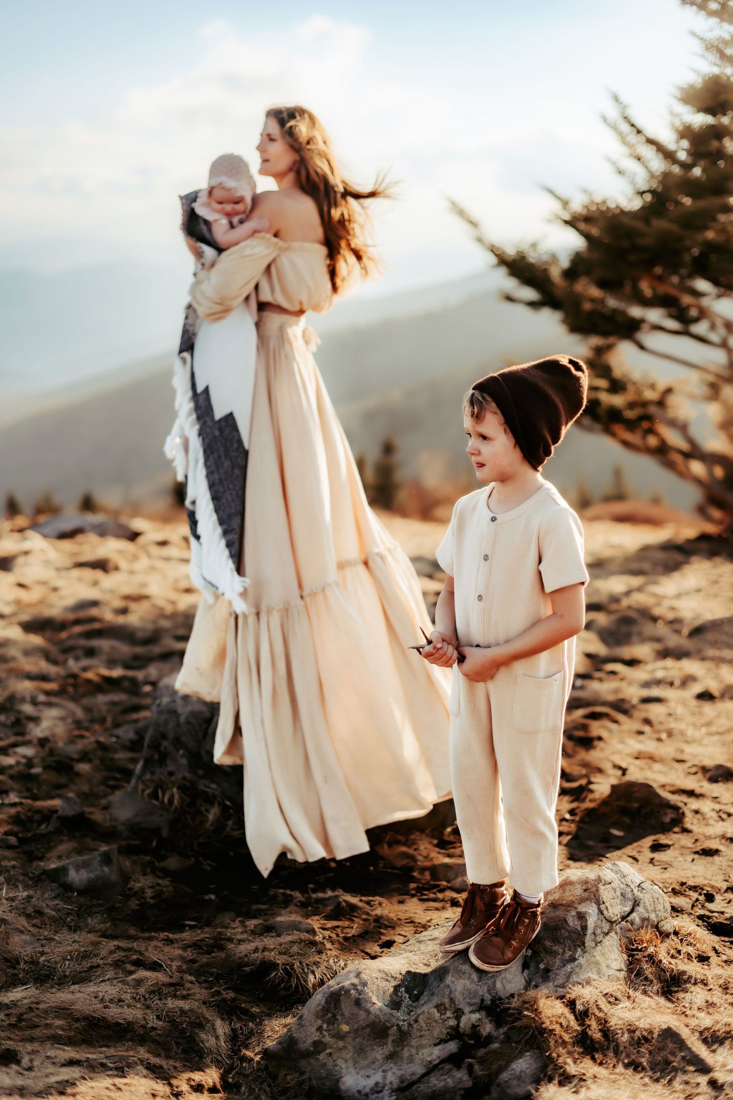 A family stands on a mountain summit at sunset with long flowing dress and casual attire against scenic views.