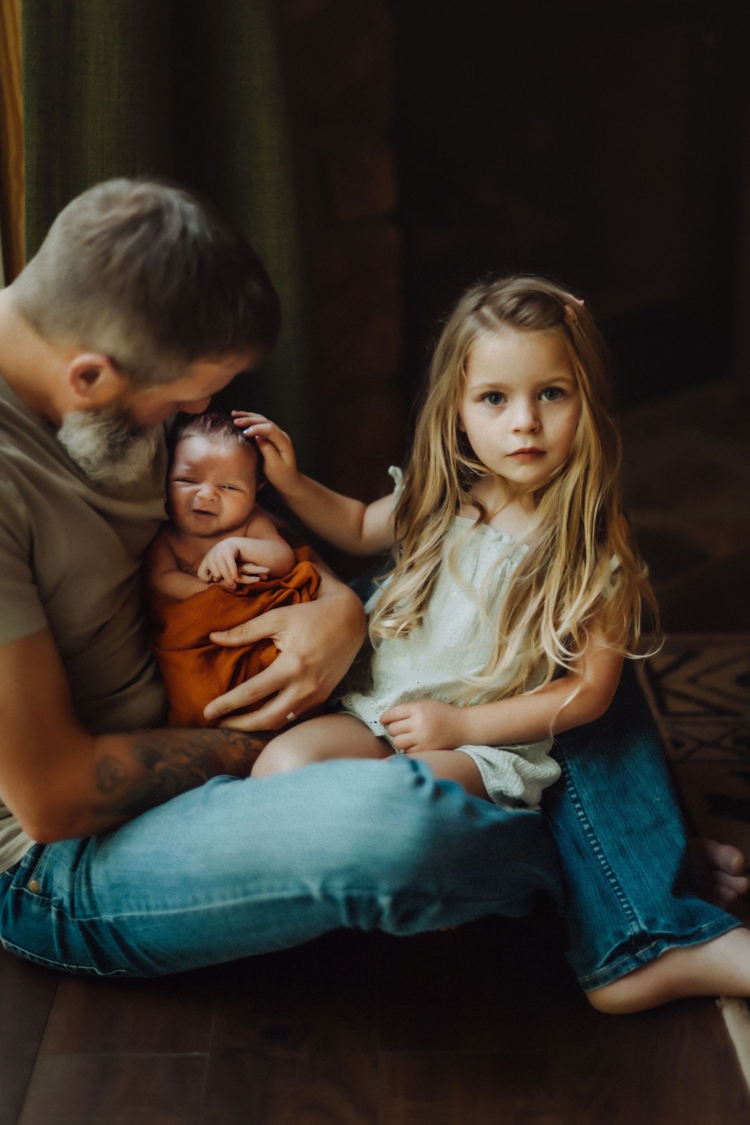 Intimate family moment captured in warm moody lighting as they sit together on wooden floor.