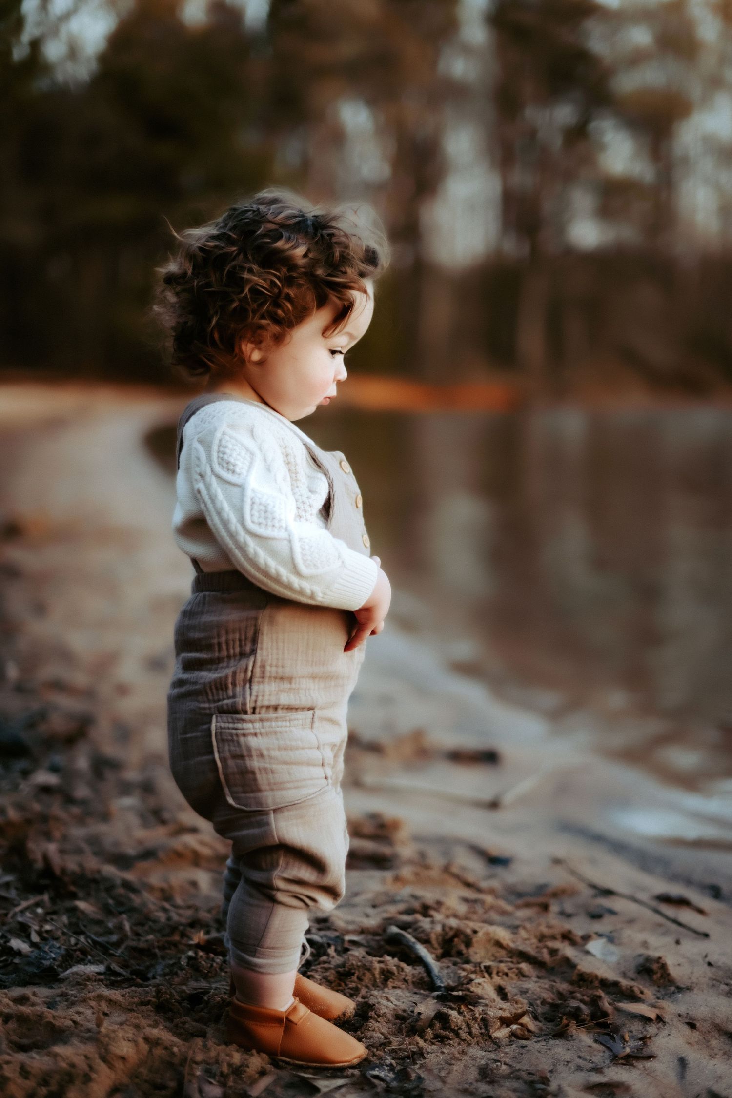 Small child in white sweater and brown pants stands thoughtfully by the water's edge at sunset.
