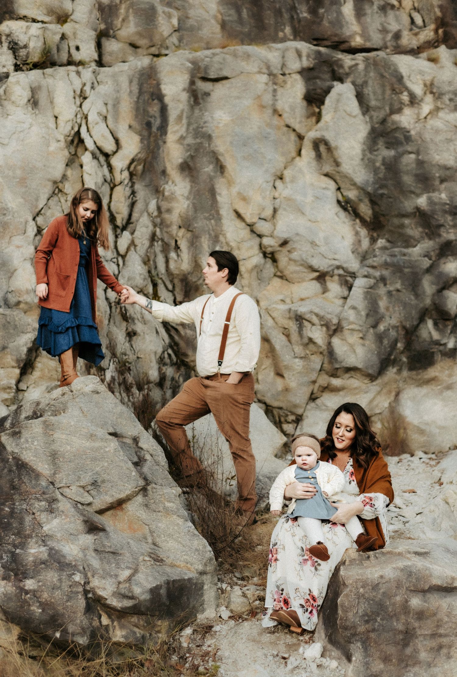 A family dressed in earth tones poses together on rocky terrain with a dramatic stone cliff face in the background.