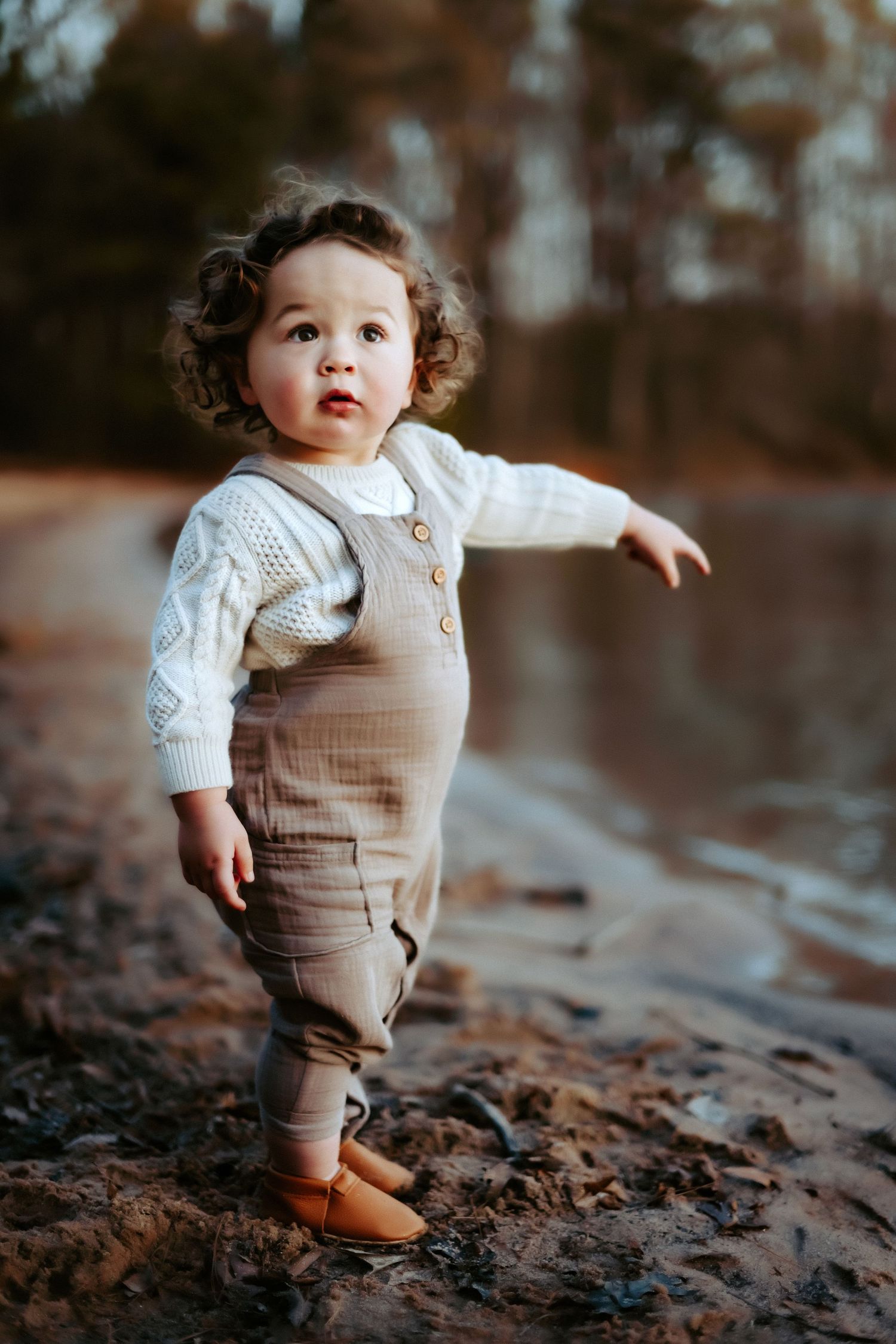 Small toddler wearing khaki overalls and white sweater stands on outdoor trail in brown leather shoes.
