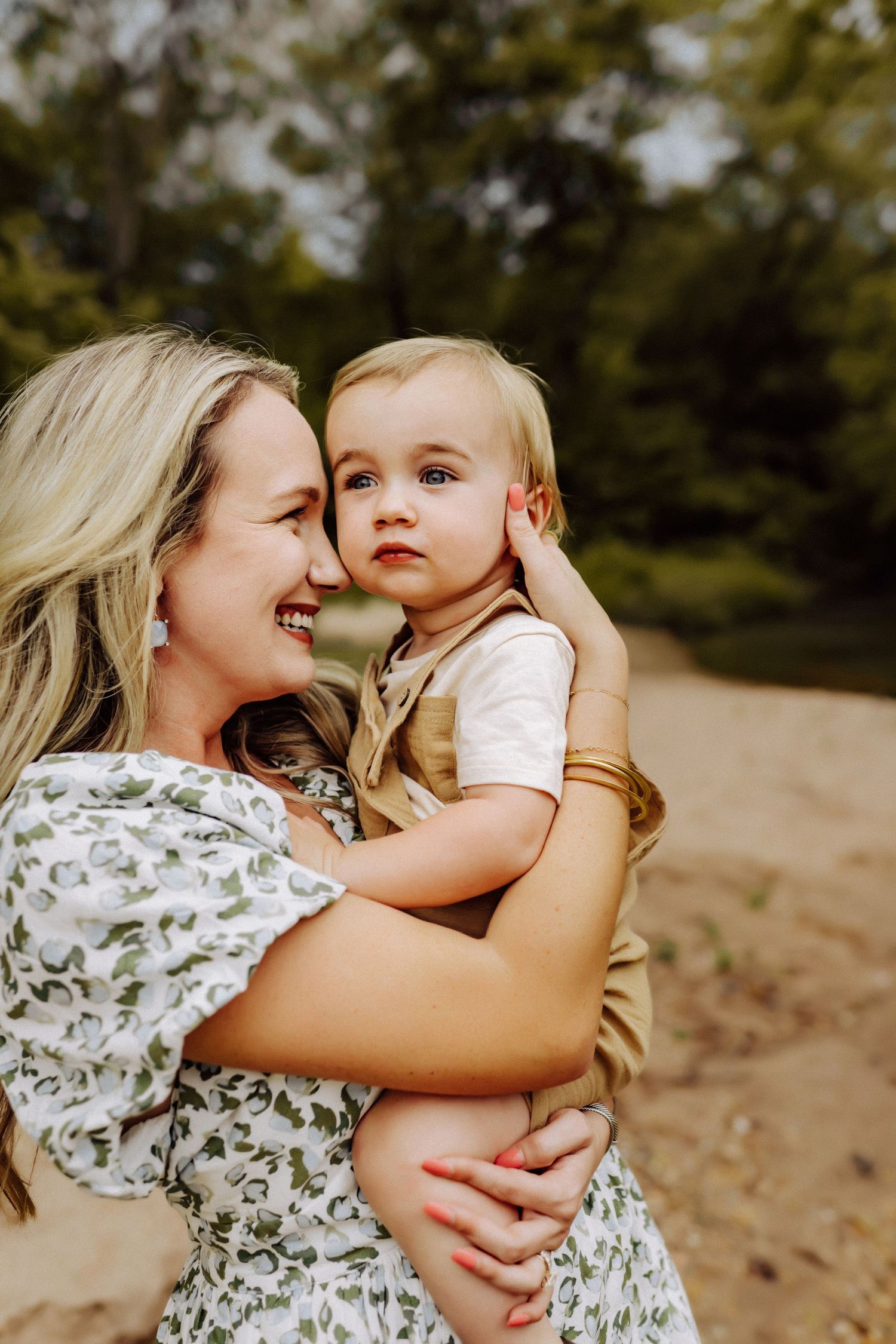 A joyful outdoor portrait of a parent in a floral dress holding a young child wearing a beige vest in nature.