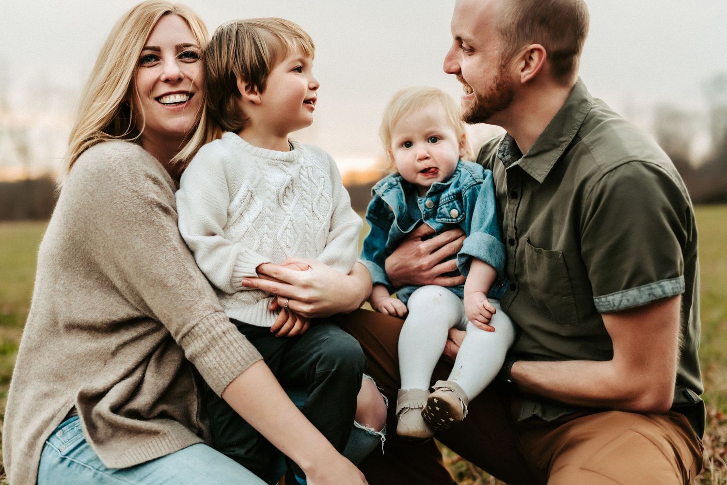 A family of four sits together outdoors, wearing casual clothing while sharing joyful expressions on a cloudy day.