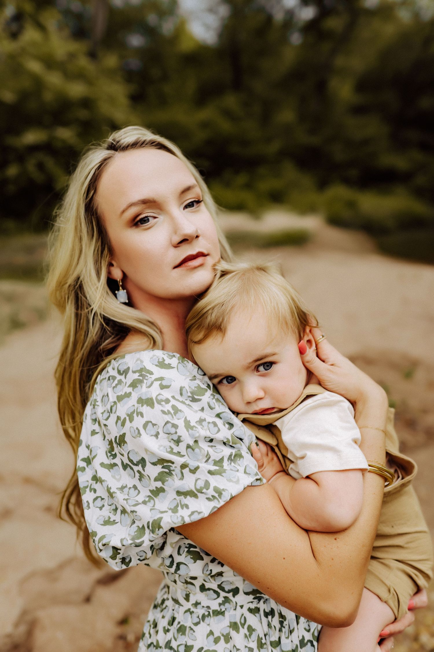 A portrait of a mother wearing a floral dress holding a baby in a light shirt on a dirt path in warm natural lighting.