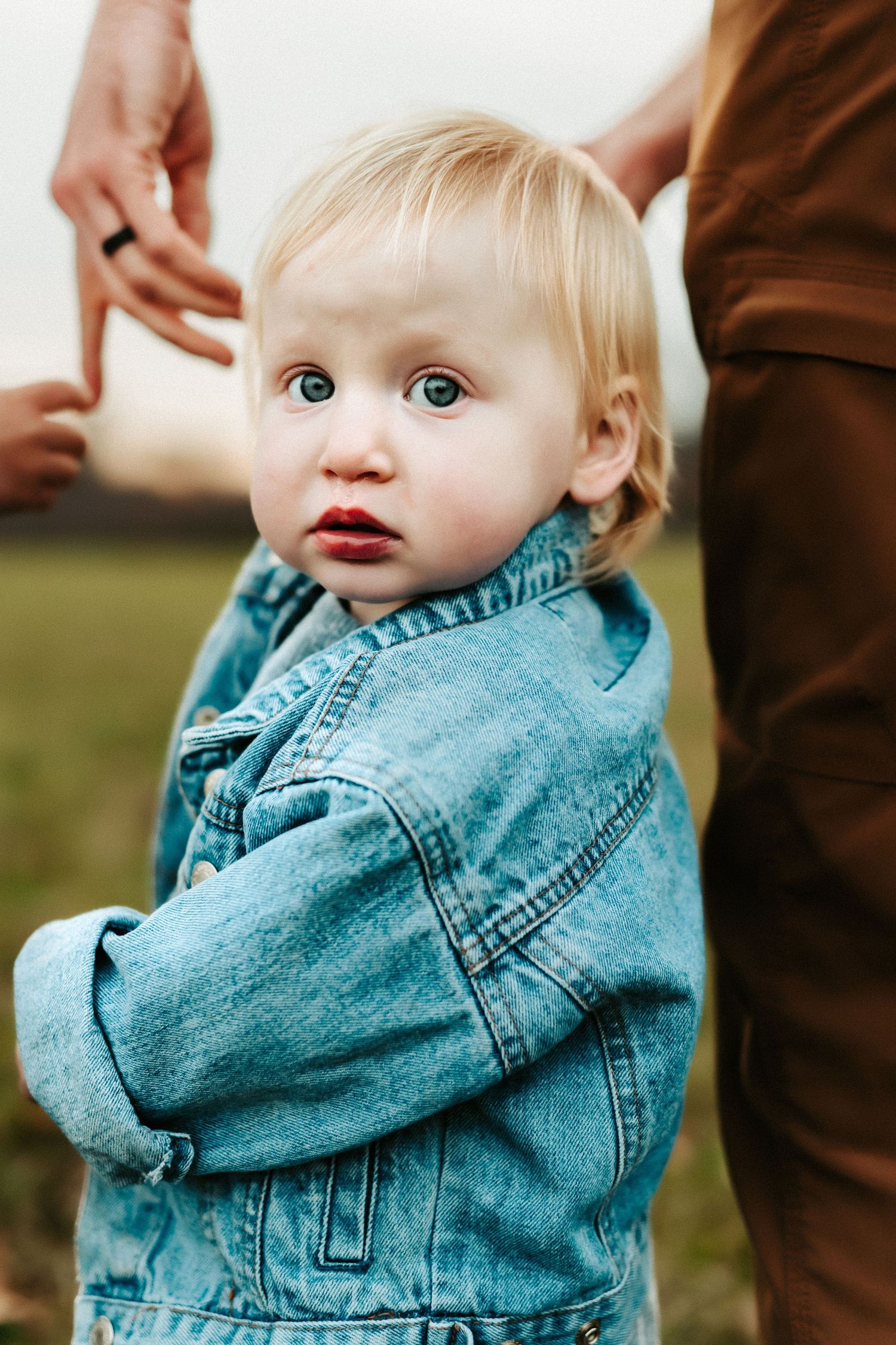 Small child wearing a denim jacket looks up with bright blue eyes while standing outdoors in green grass.