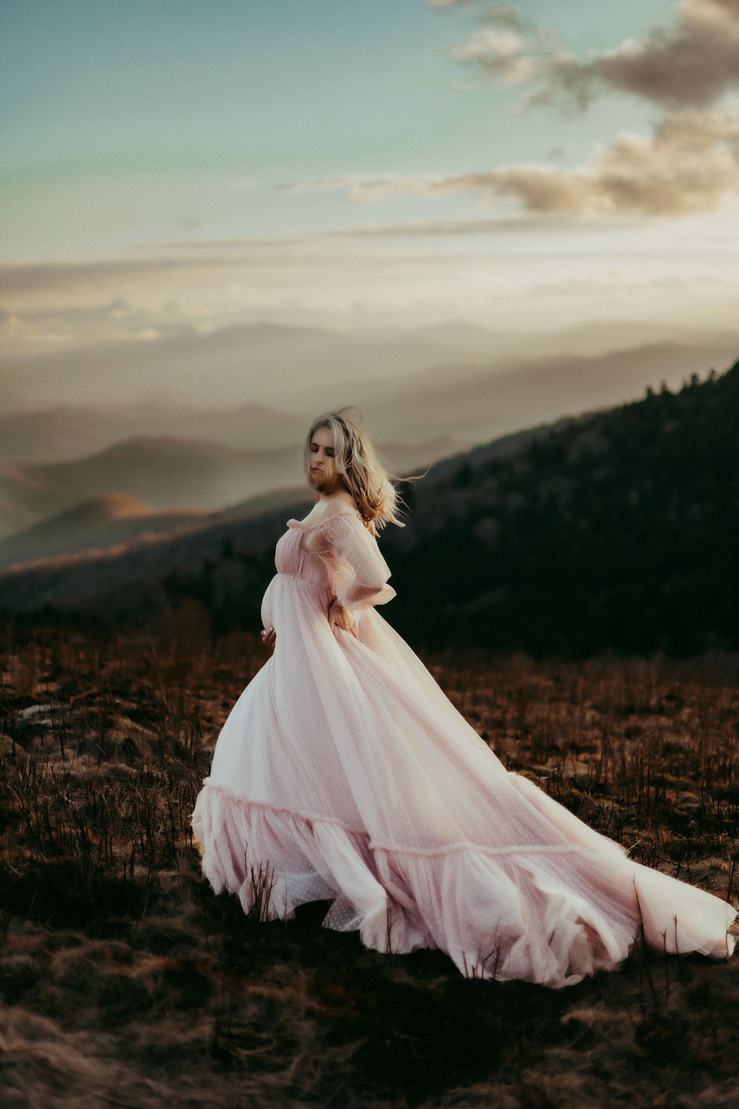 A flowing pink gown billows in the wind against a misty mountain backdrop during golden hour sunset.