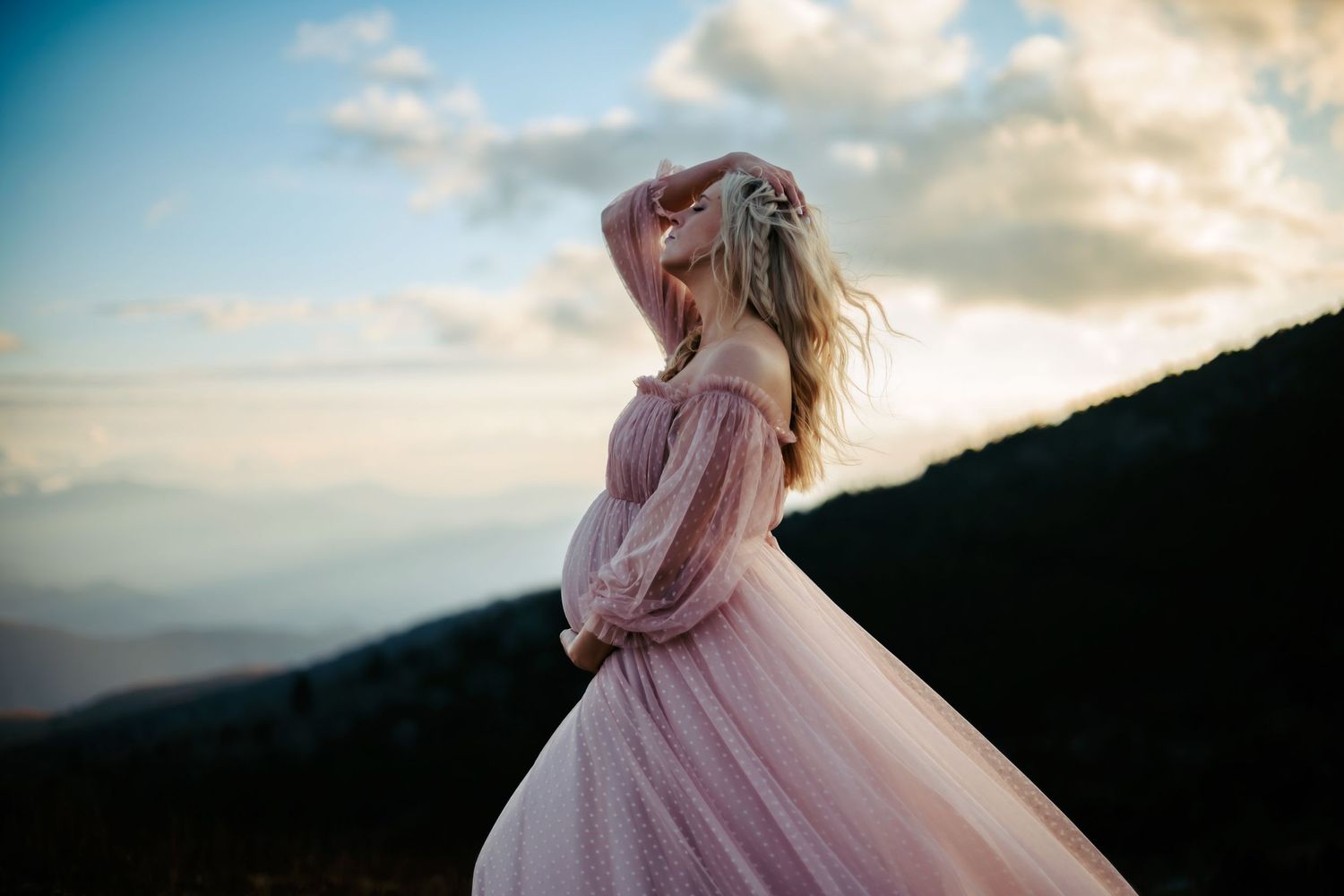 A woman in a flowing pink dress stands on a mountaintop silhouetted against a cloudy sunset sky.