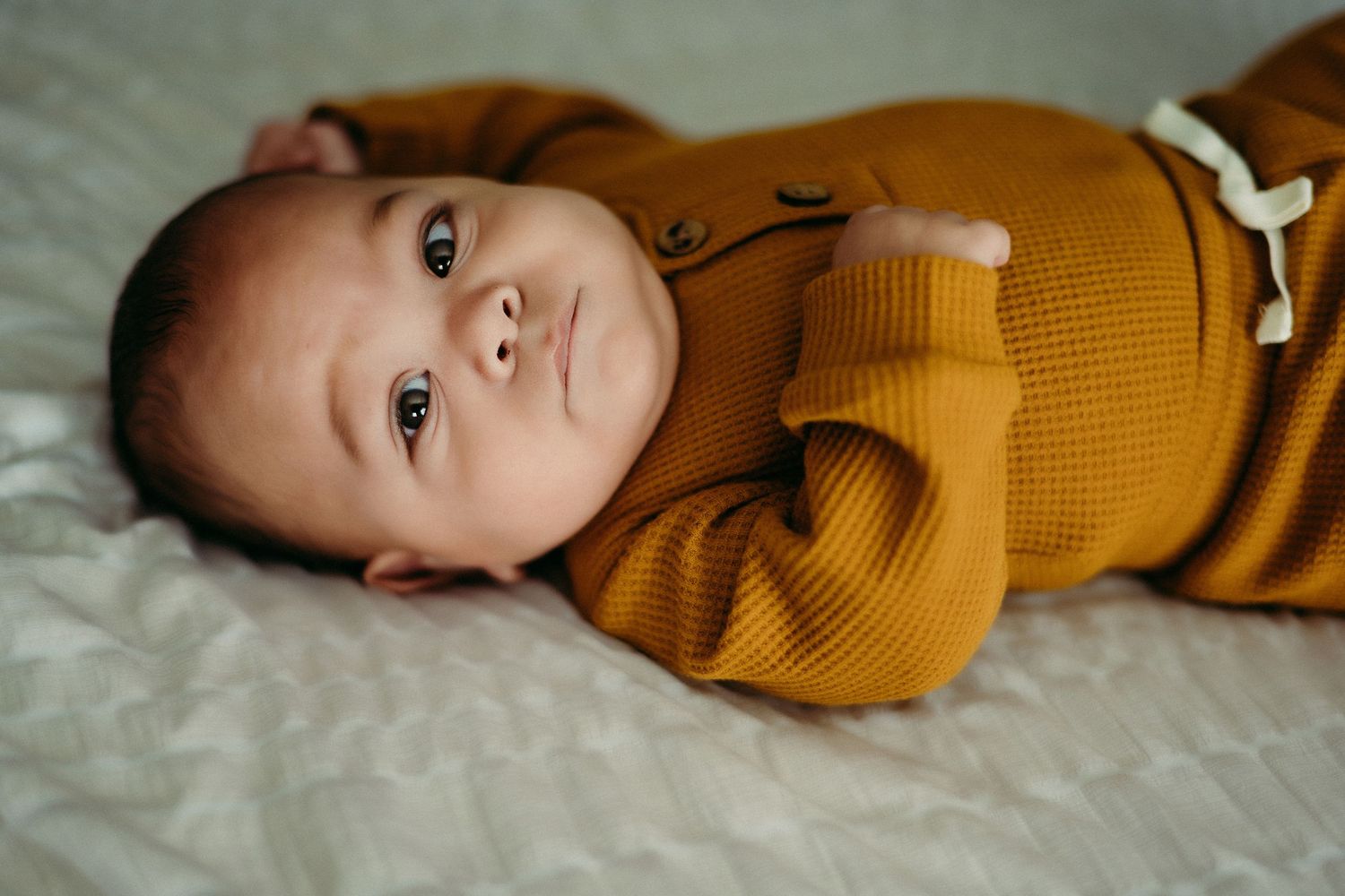 Baby wearing a cozy mustard yellow sweater lies on soft white bedding.