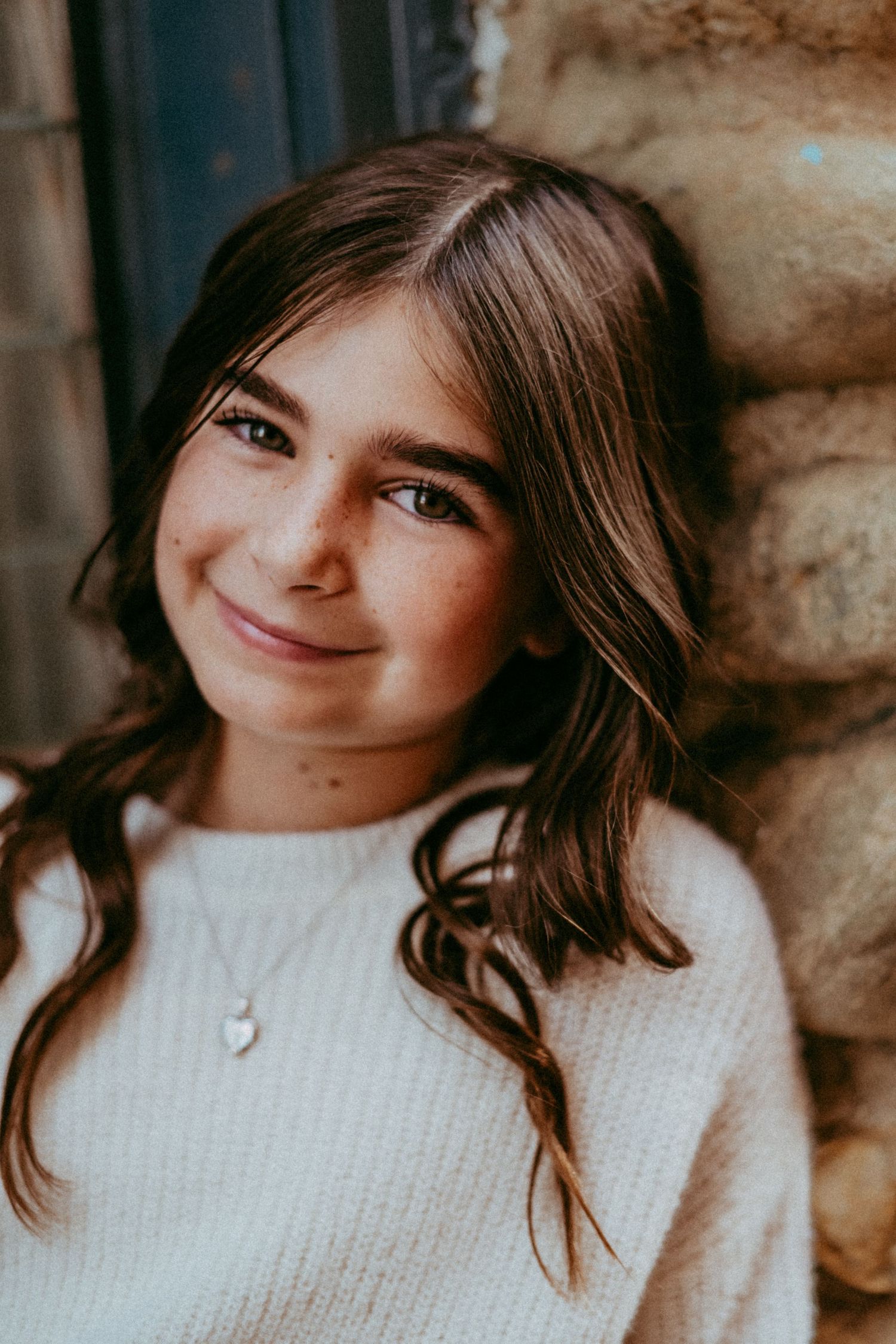 Young person with long brown hair and warm smile wearing white knit sweater and necklace against stone wall background.