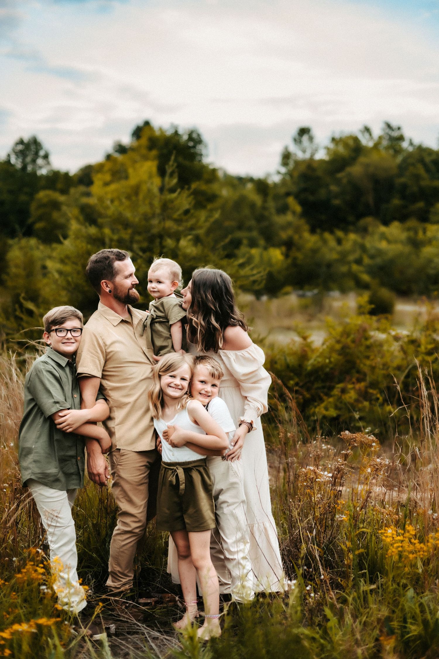 A family stands together in a golden meadow during autumn with trees and cloudy skies in the background.