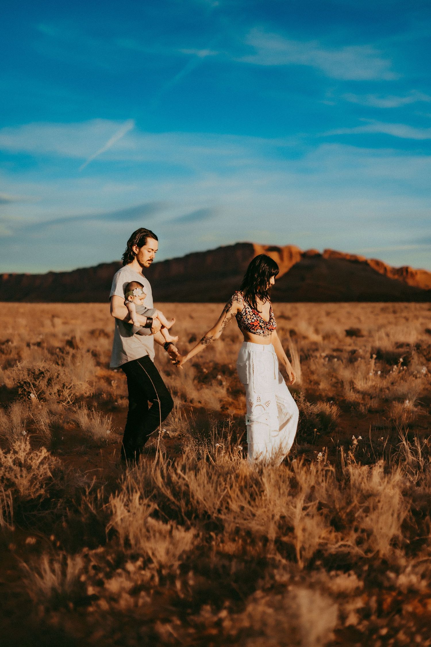A couple walks hand in hand through a golden desert field at sunset with mountains in the background.