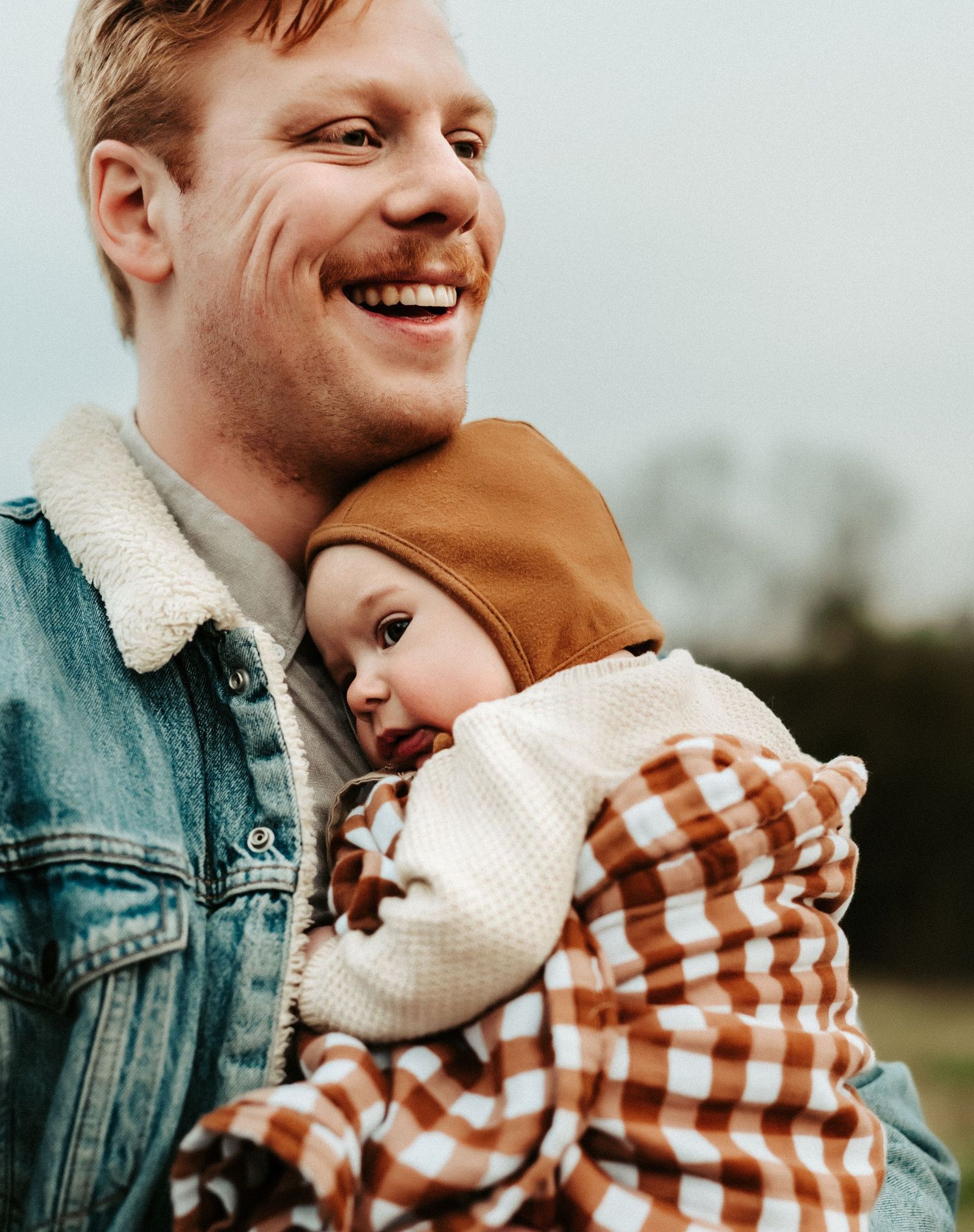 A tender parental moment captured outdoors with a baby wearing a plaid outfit and white sweater.