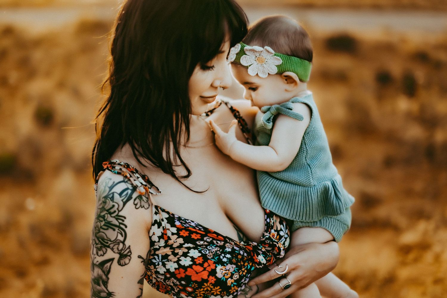 A woman in a floral dress holds a baby wearing a mint green sweater against a golden autumn background.