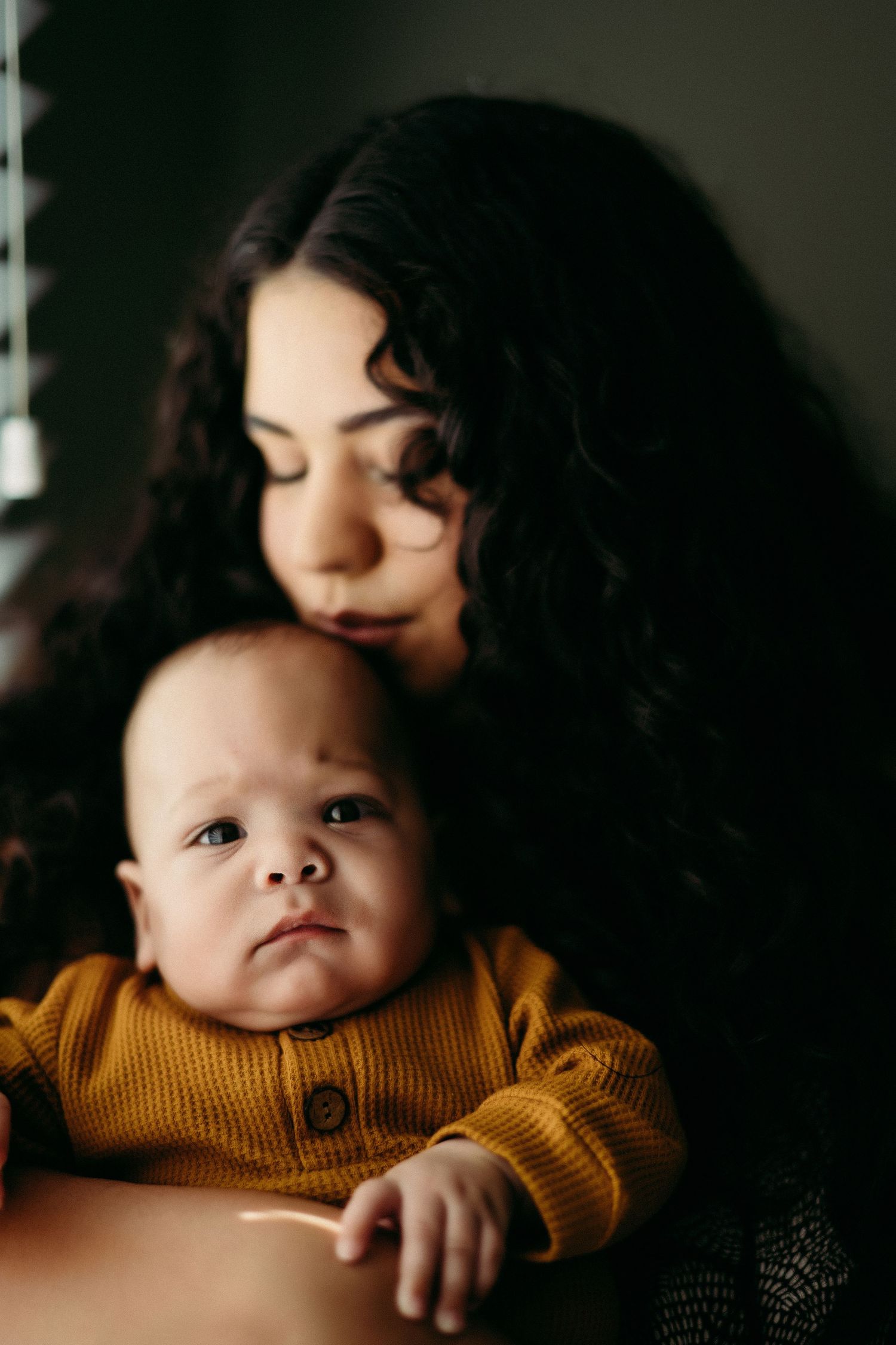 An intimate portrait shows a parent embracing a baby wearing a mustard yellow sweater in moody lighting.