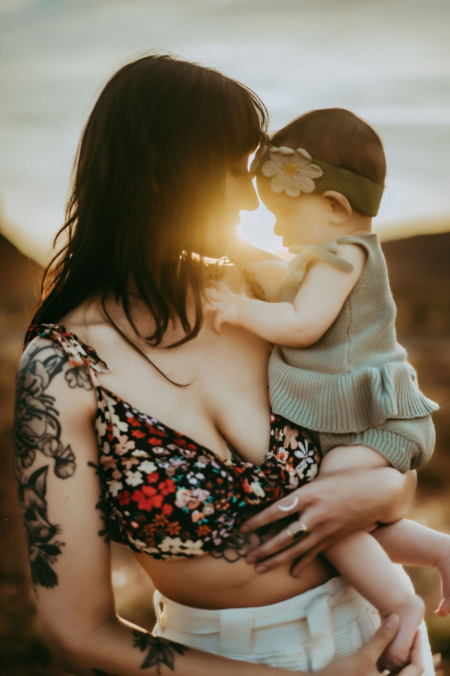 Mother in floral top with tattoo holds infant in grey outfit during golden sunset.