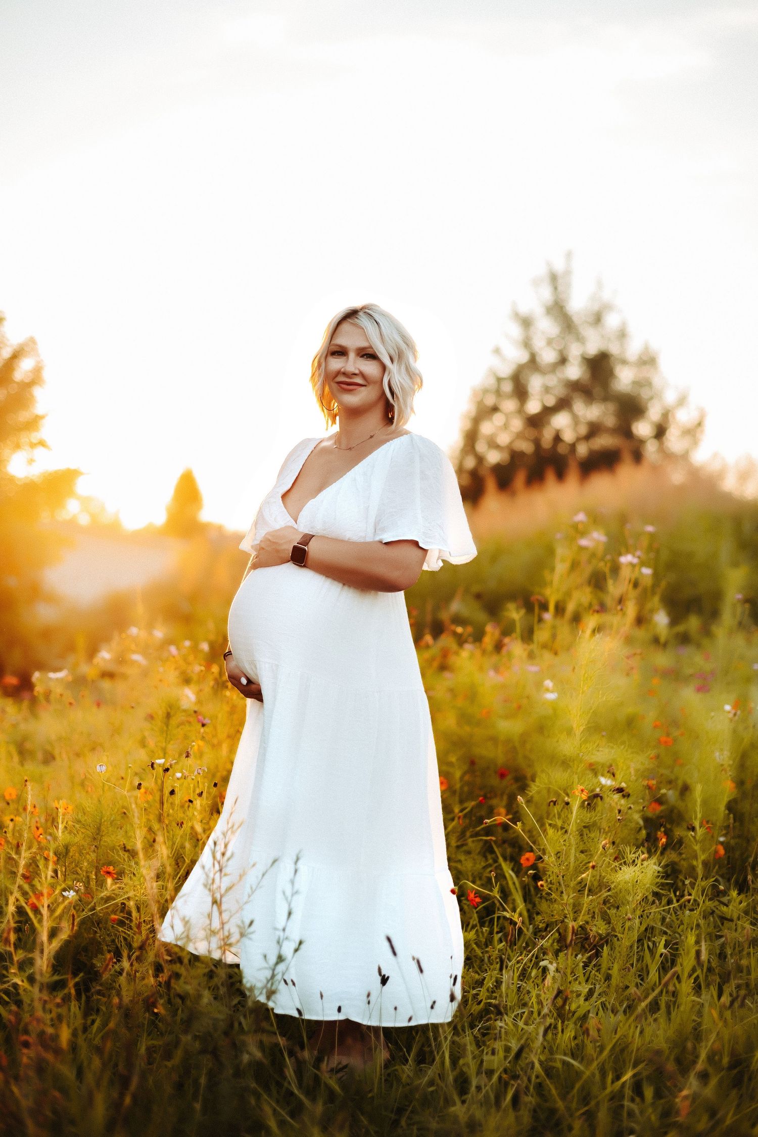 A pregnant woman in a white maxi dress stands in a golden sunlit field at sunset, surrounded by wildflowers.