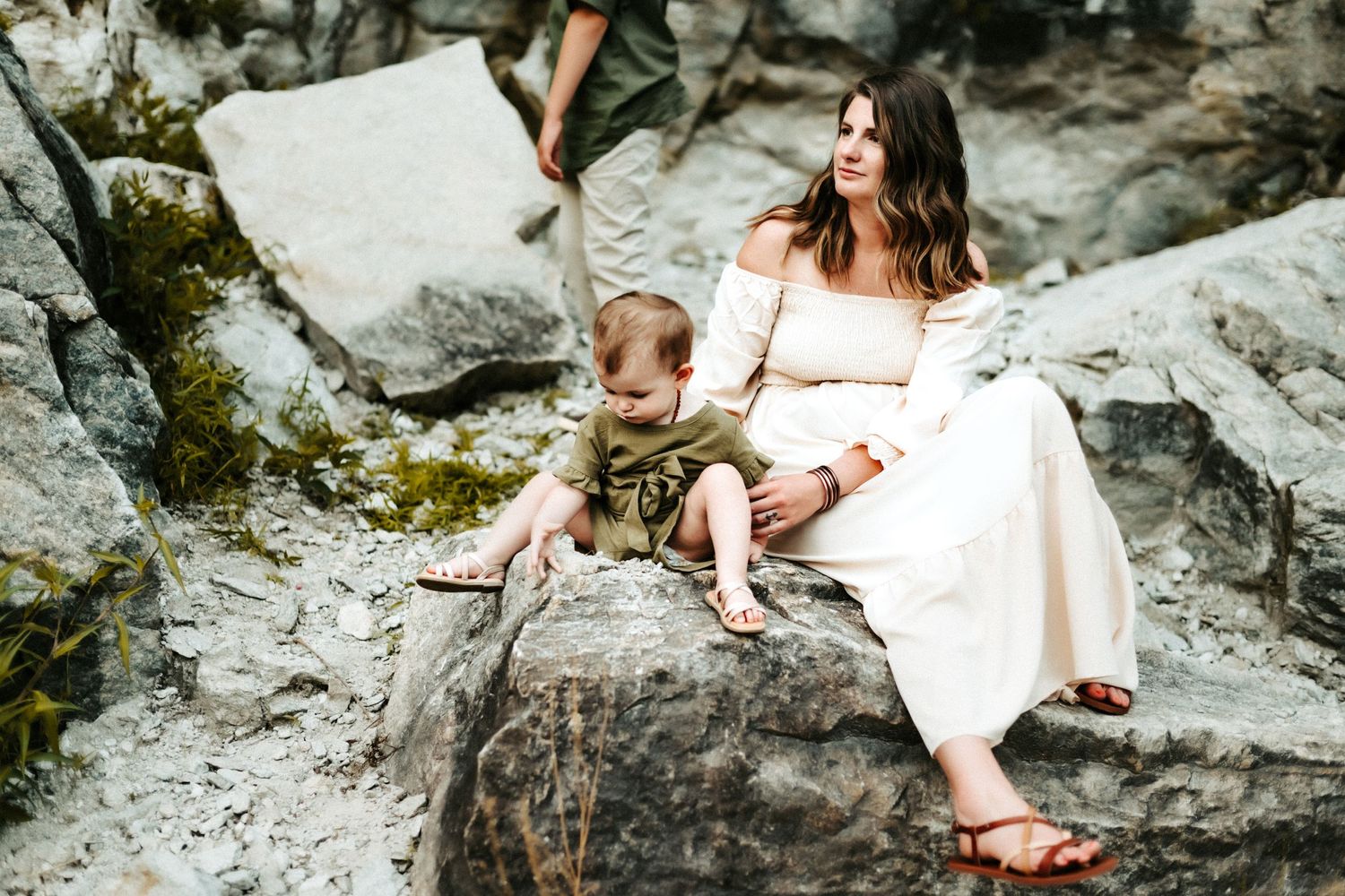 A woman in a white dress sits with a child on moss-covered rocks near water.