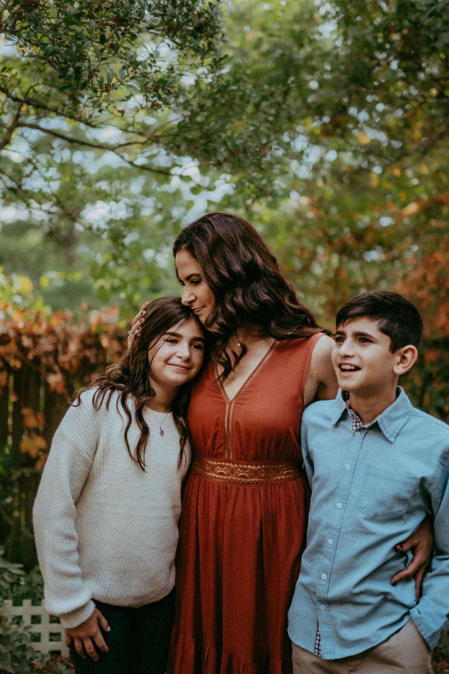 A family portrait outdoors with three people smiling together in front of greenery and autumn colors.