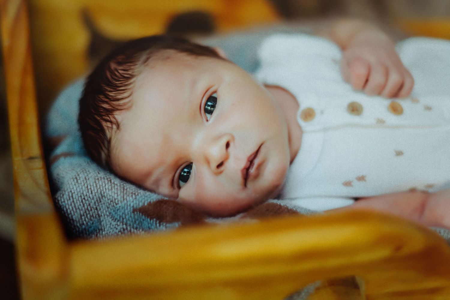 A baby wearing a white onesie lies on blue fabric against a blurred golden yellow background.