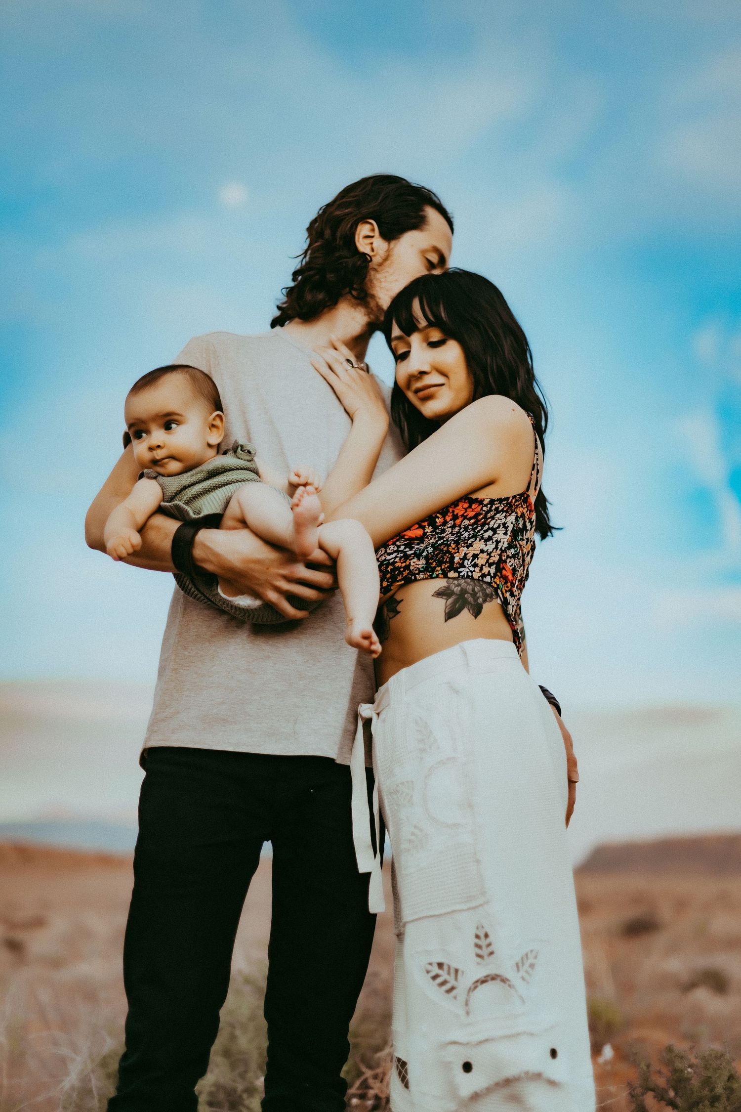 A couple embraces while holding a baby against a blue sky and desert landscape background.