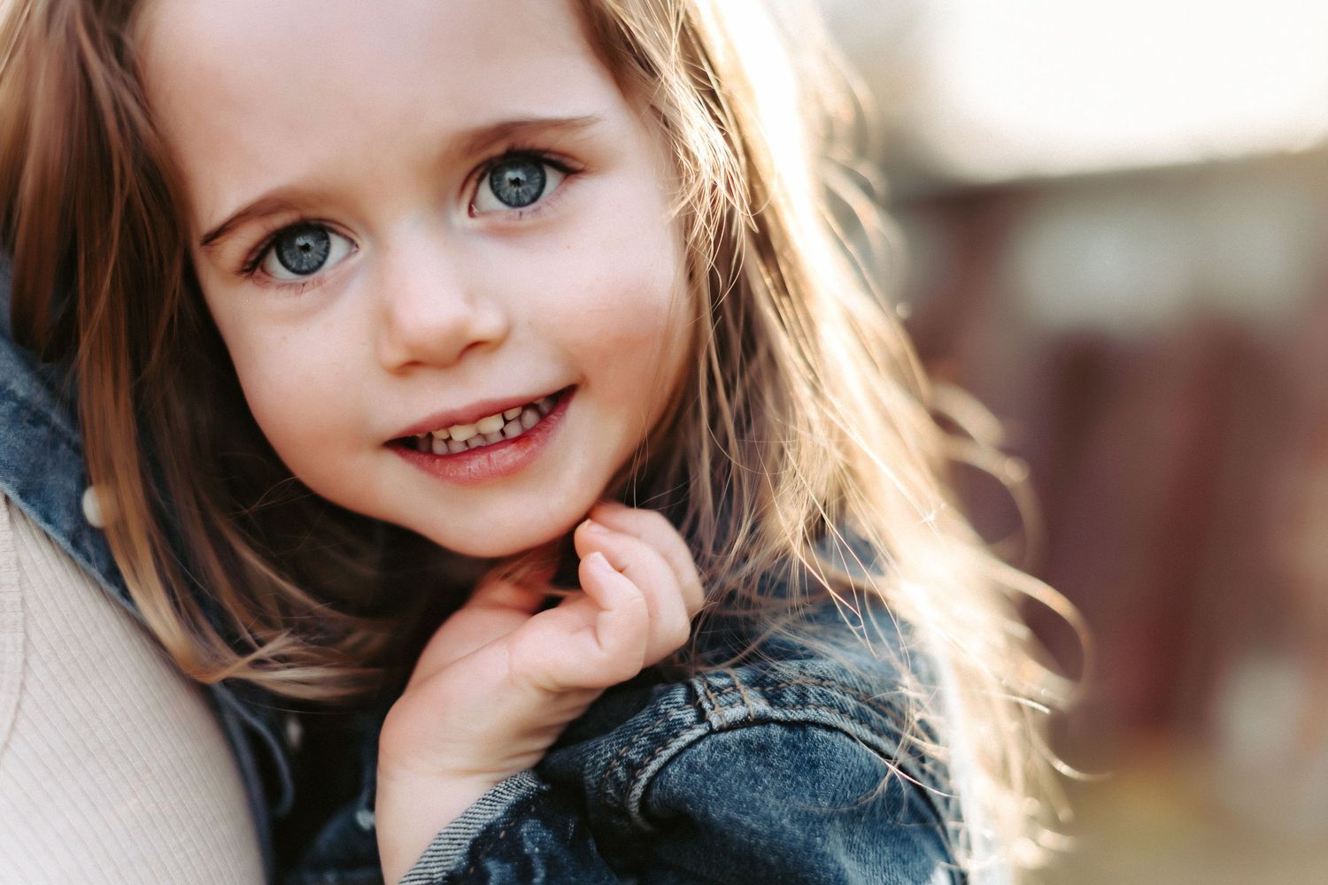 Child in denim jacket smiles while resting against a light surface in soft lighting.