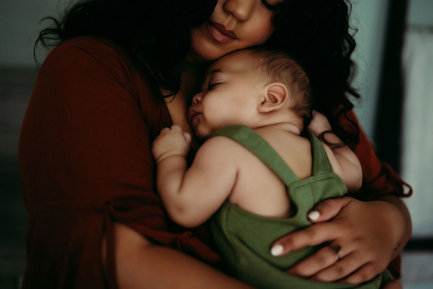 A sleeping baby in a green romper rests peacefully against a caregiver in a dark maroon sweater.