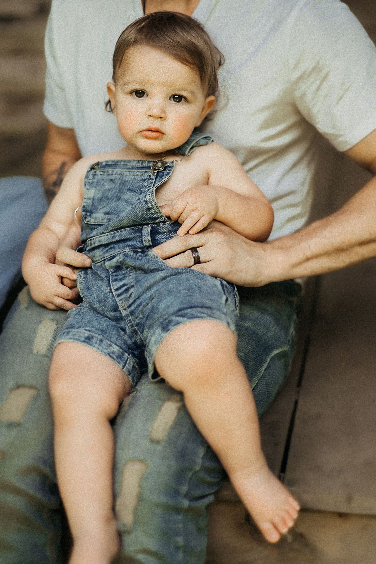Small child wearing blue jean overalls sits on a caregiver's lap with bare feet.