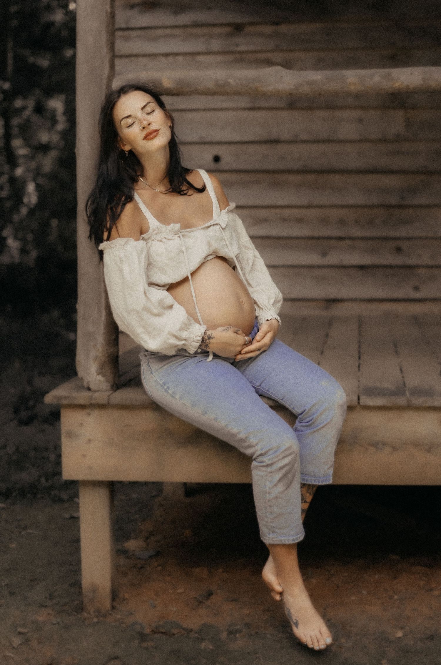 Pregnant person in white peasant top and gray pants sits barefoot on wooden porch during maternity photoshoot.