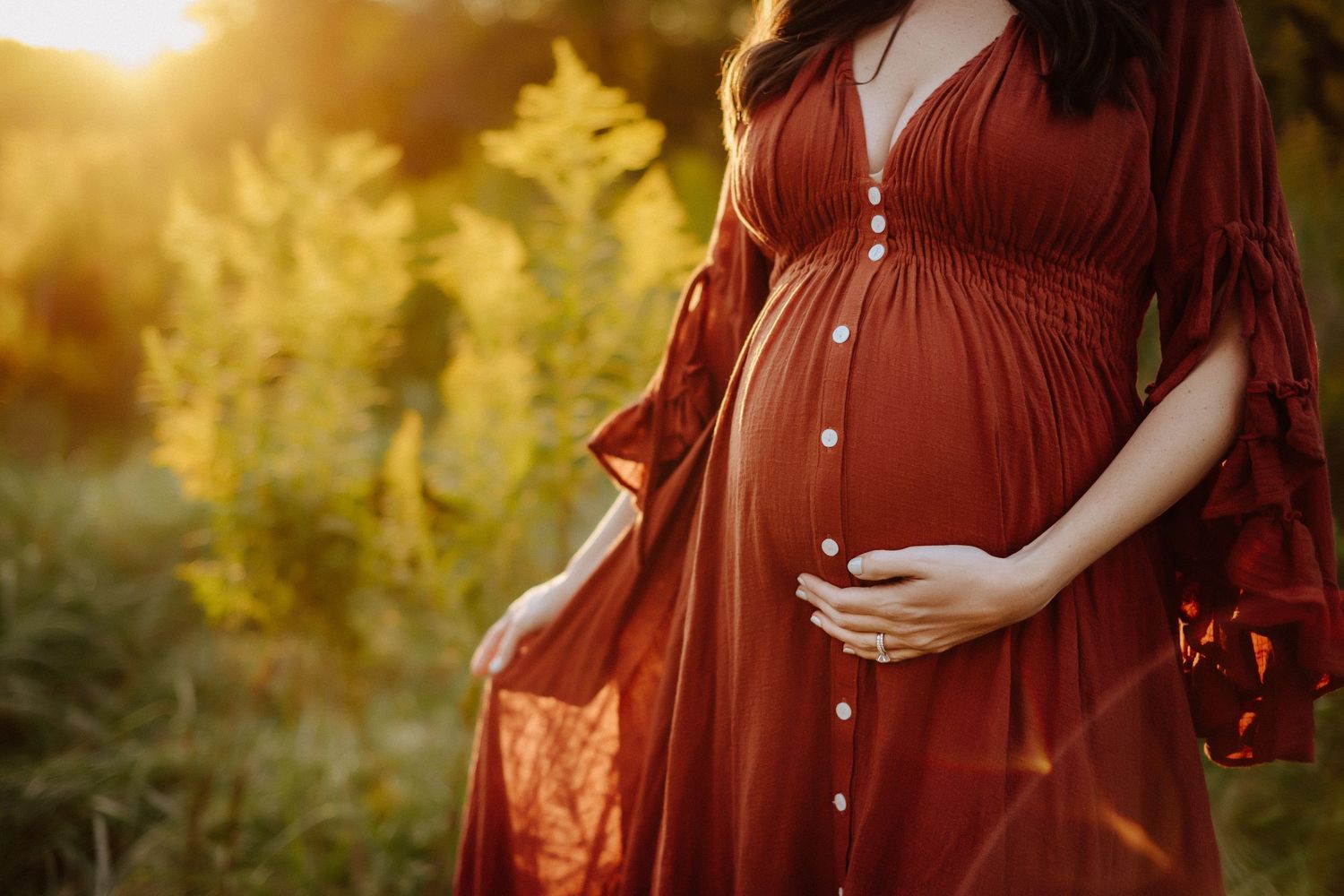 Pregnant woman in flowing burgundy dress cradles belly during golden hour sunset photoshoot in nature.