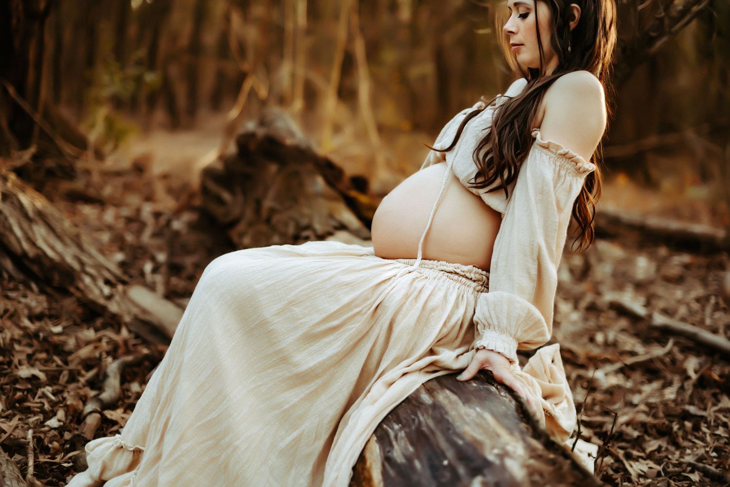 A pregnant woman in a flowing white dress poses in a serene autumn forest setting during golden hour.